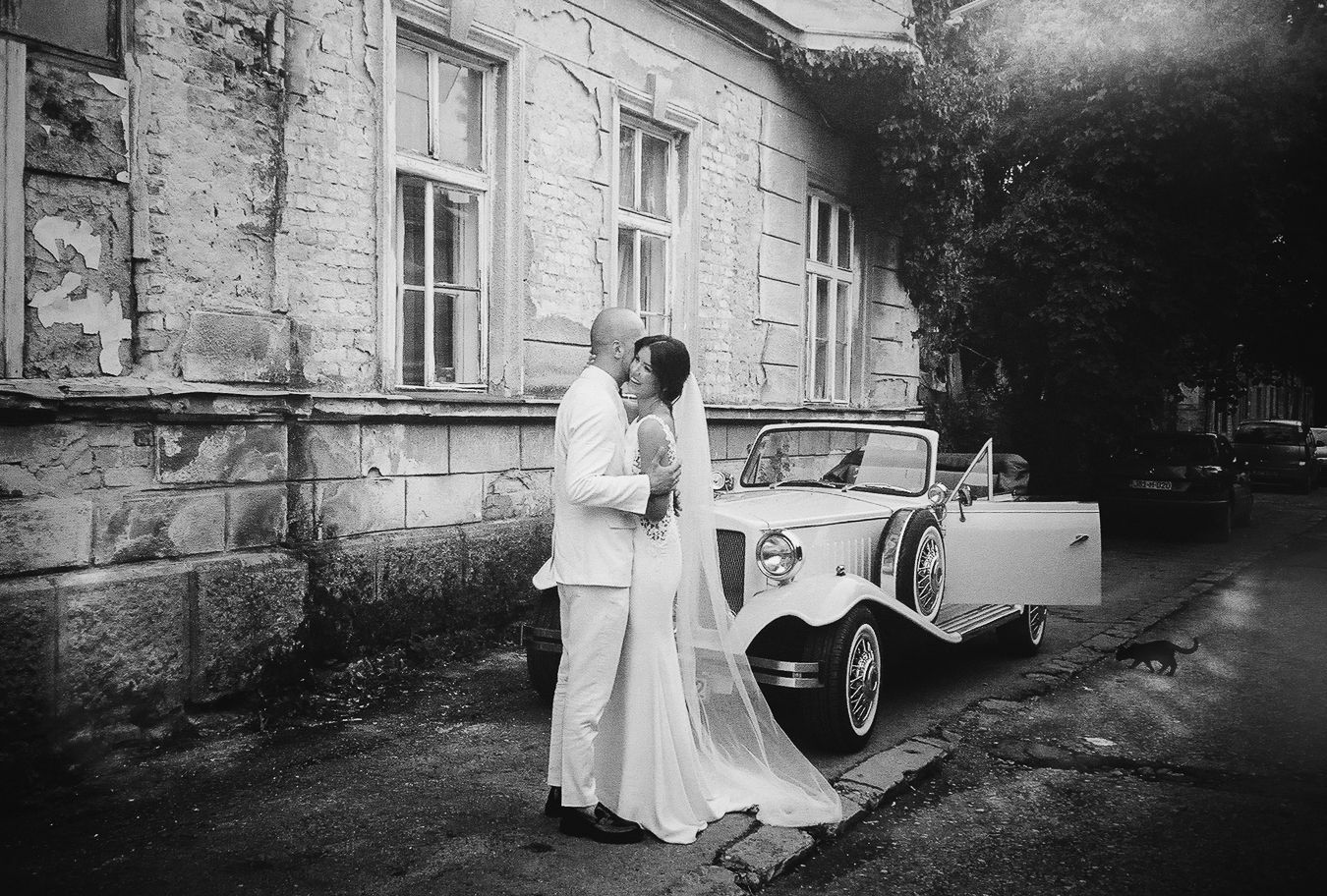Black and white wedding photo of bride and groom kissing beside a vintage white car outside an old stone building.