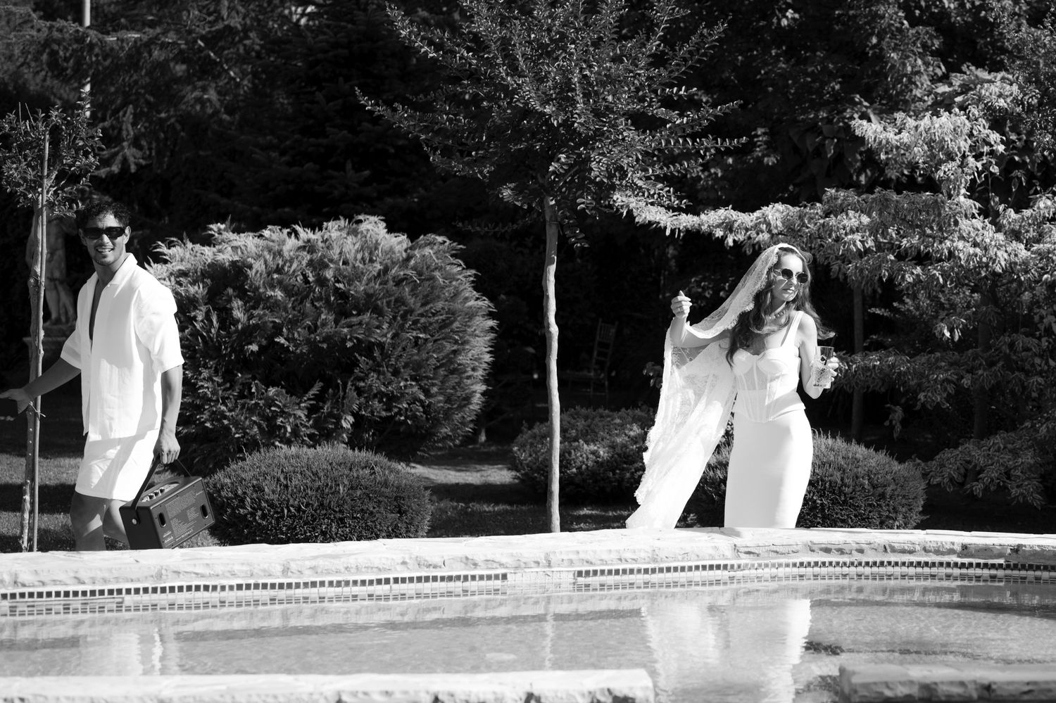 Crno-bijela fotografija mladenaca u bijelim autfitima pored fontane u bašti, pool party vjenčanje Villa Slatina Banja Luka