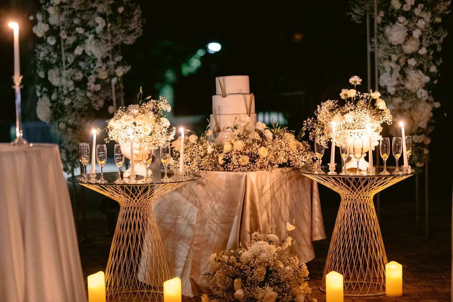 Elegant wedding cake table with white floral arrangements, candles, and gold geometric stands in a romantic outdoor setting.