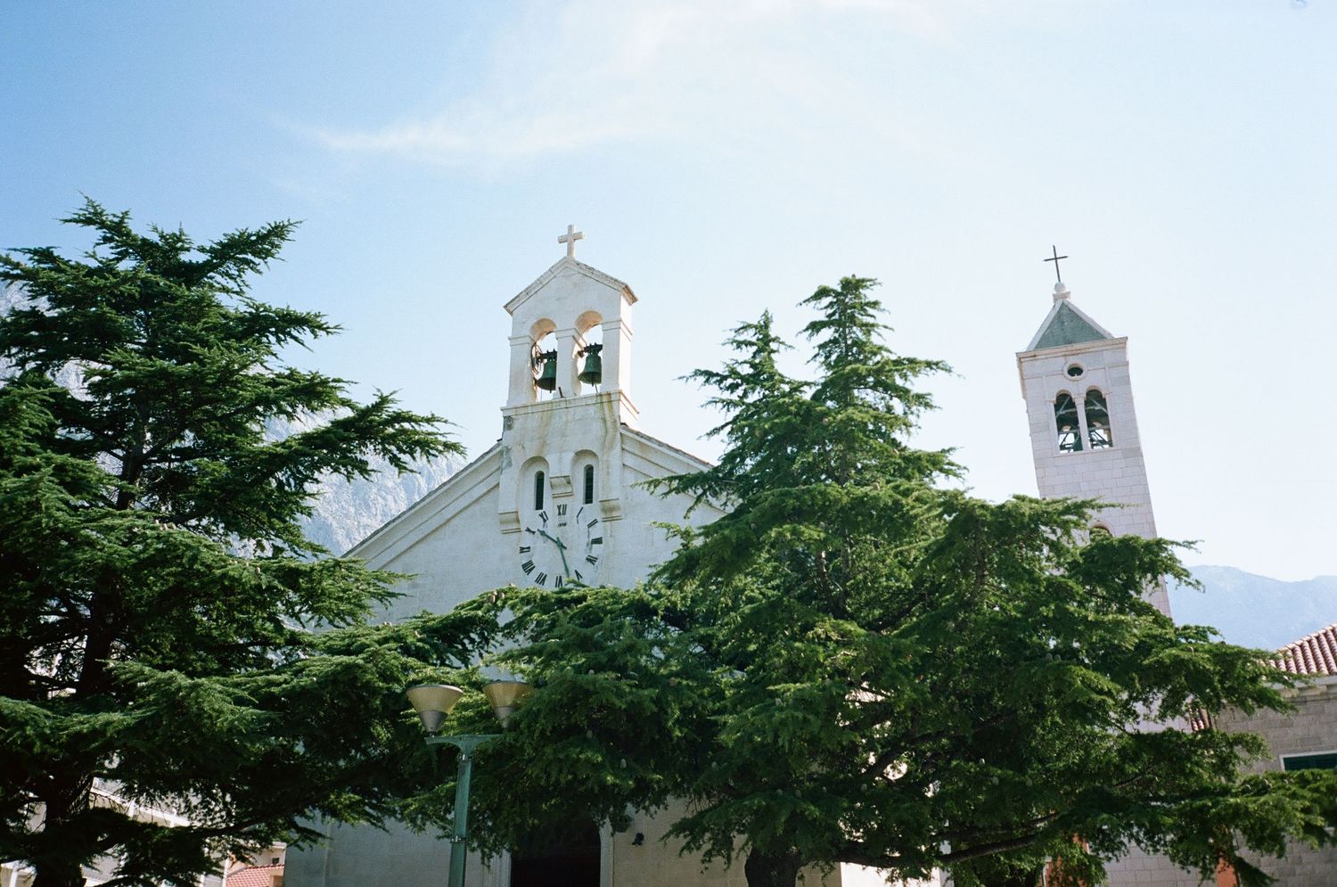 White church steeples rise above evergreen trees against a bright blue sky in a Mediterranean coastal setting.