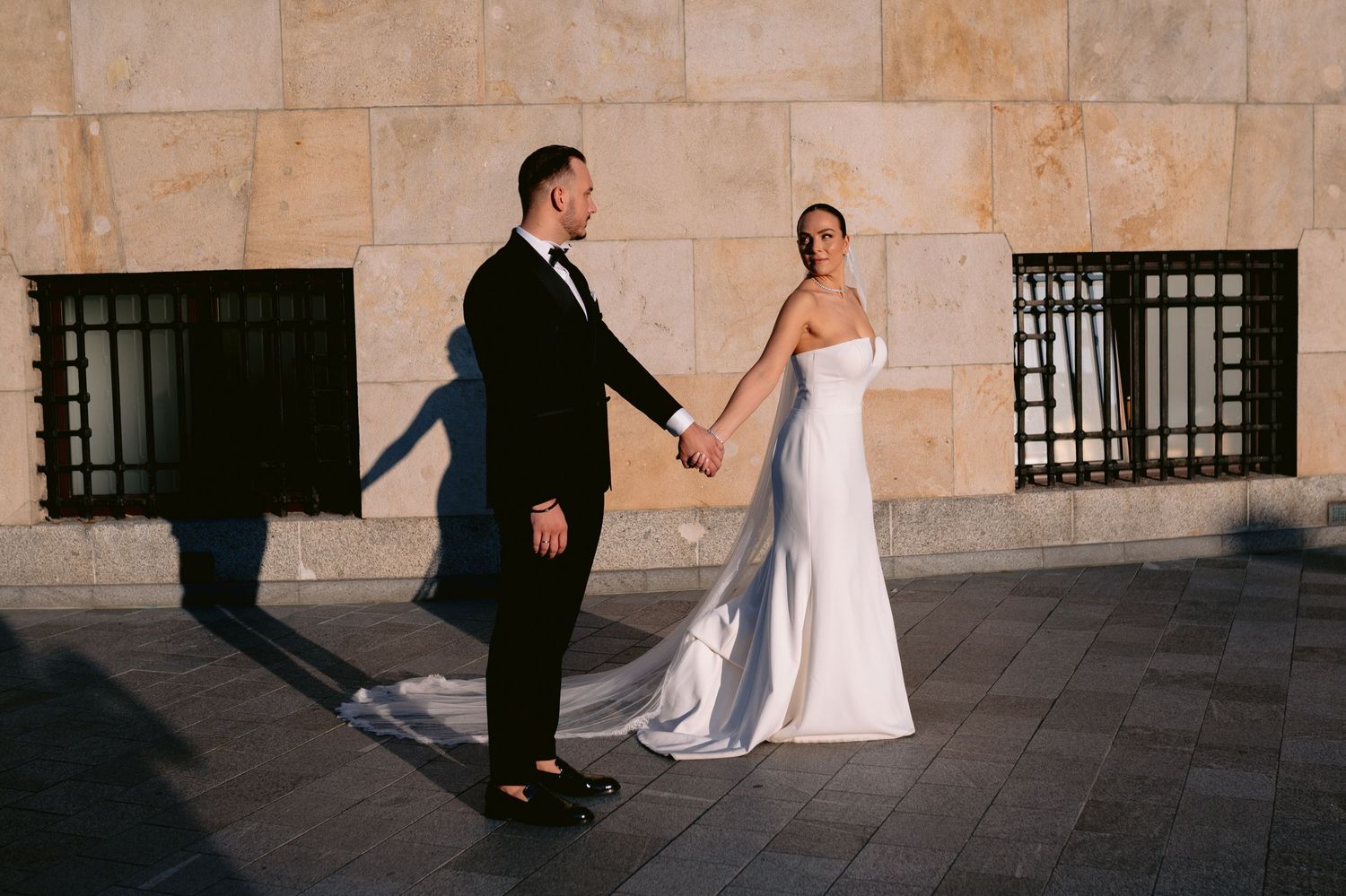 A couple in wedding attire poses for artistic photos in a stone corridor with dramatic lighting and shadows.