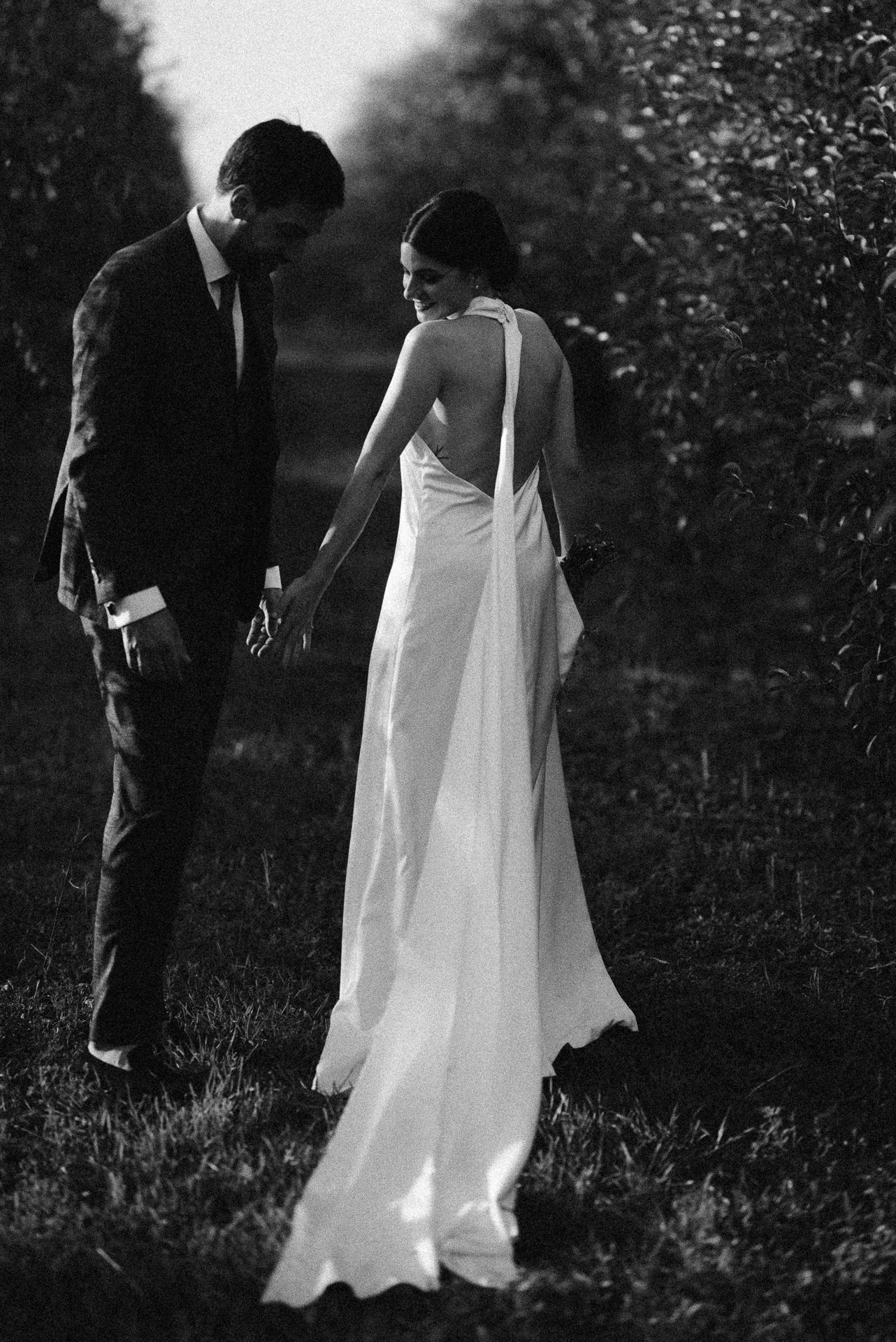 Black and white photo of a bride in backless gown and groom in suit holding hands in a lush garden setting.