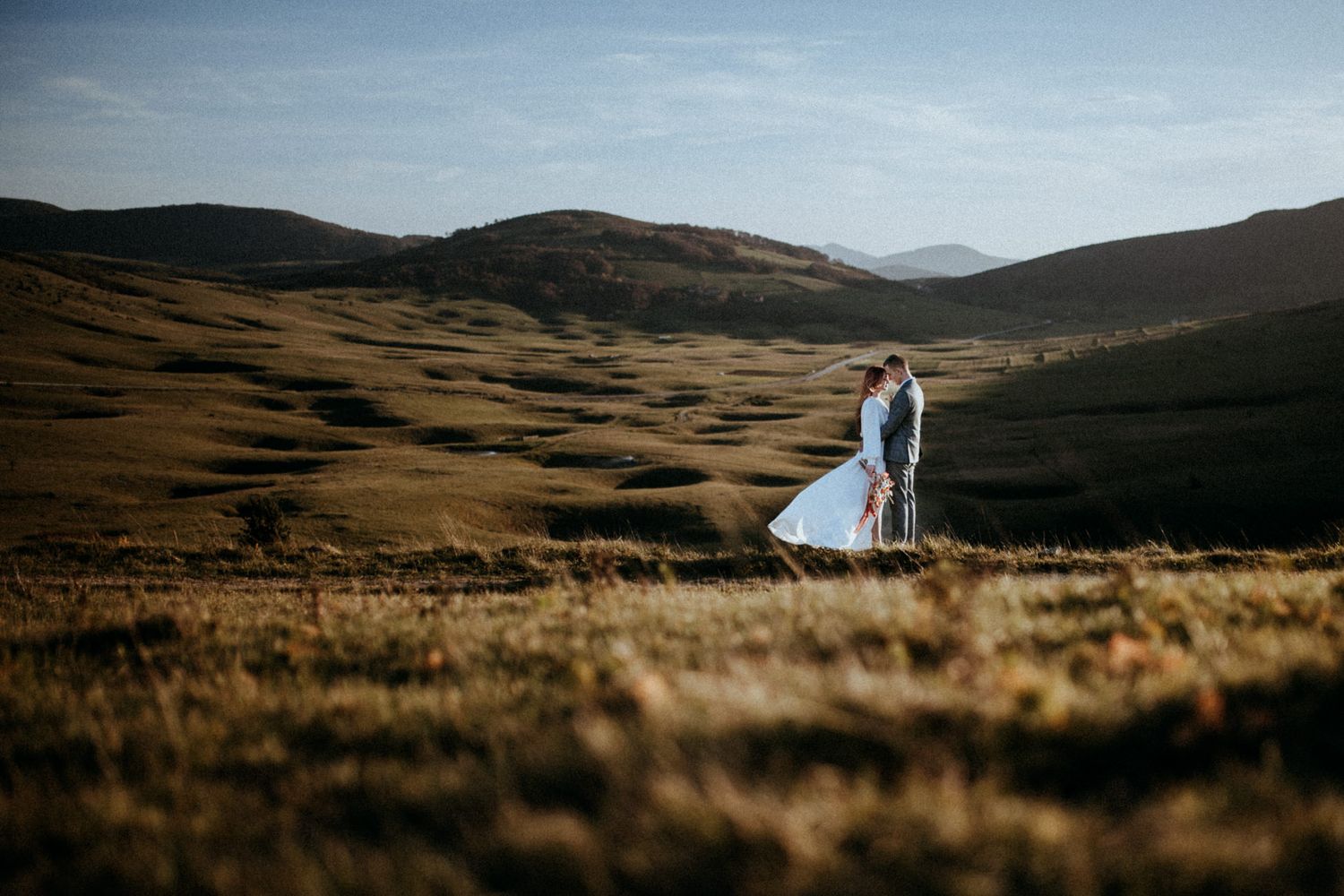 Couple sharing a romantic kiss in a vast golden meadow with rolling hills and mountains in the background.