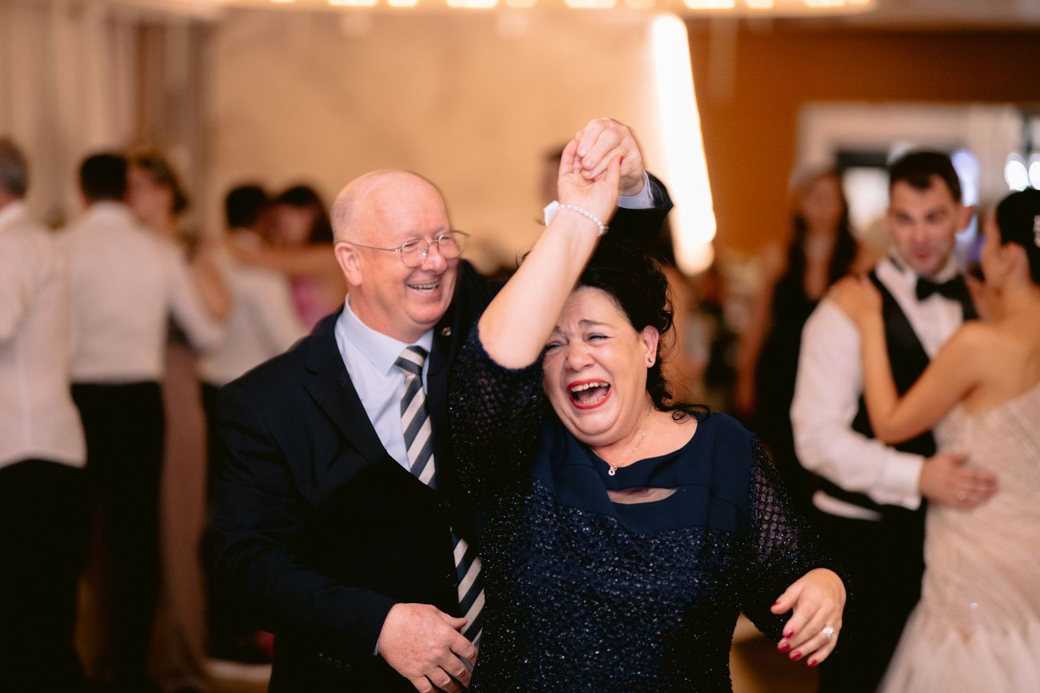 Joyful couple dancing at wedding reception, woman raises arm triumphantly in navy sequin dress on crowded dance floor.