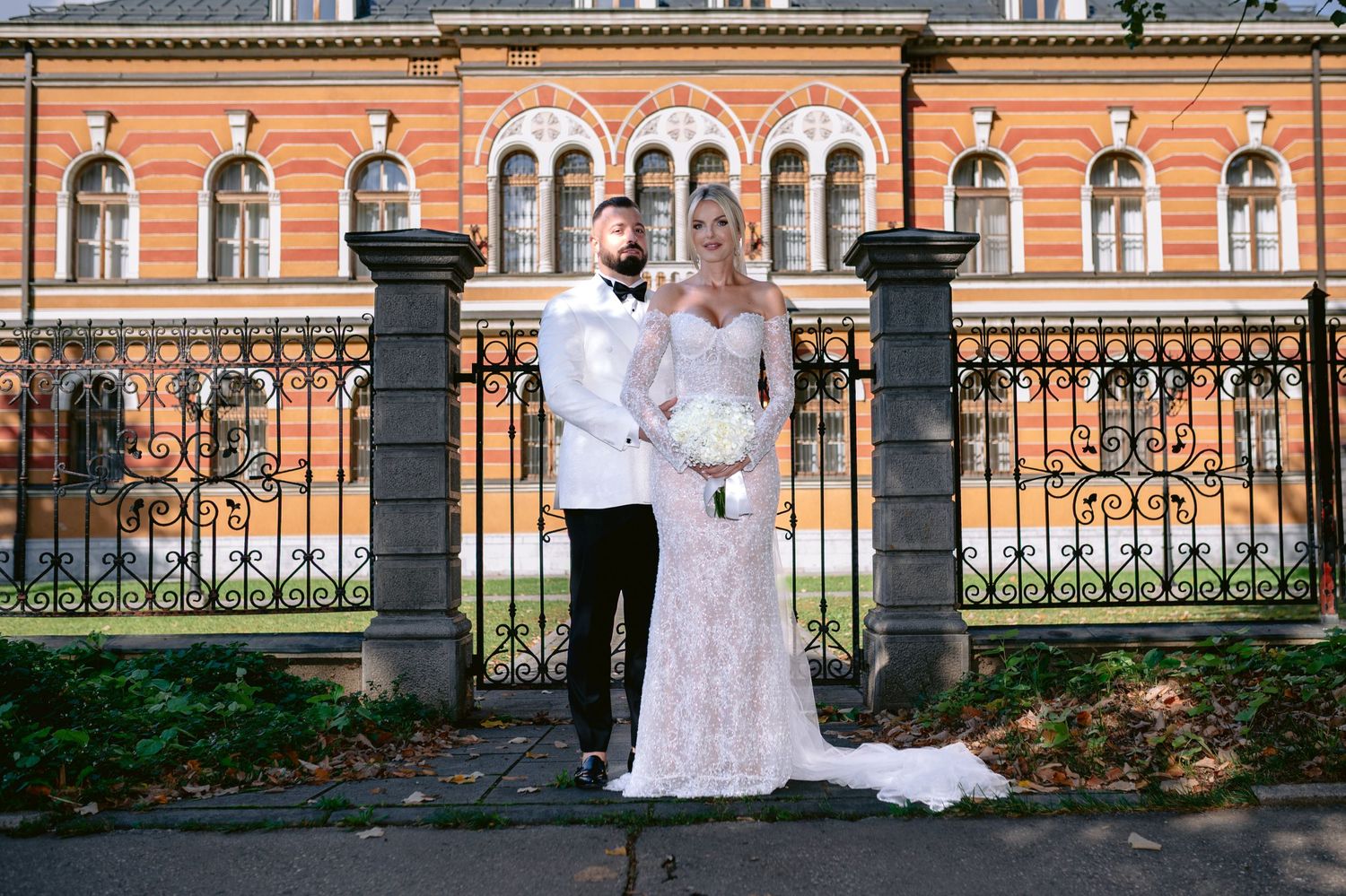 Wedding couple stands in formal attire before ornate historic building with iron fence.