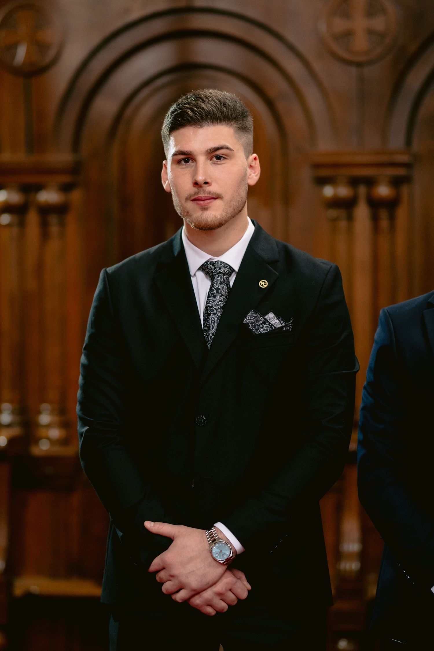A person in a black suit and tie stands in an elegant church or cathedral with ornate wooden architecture.