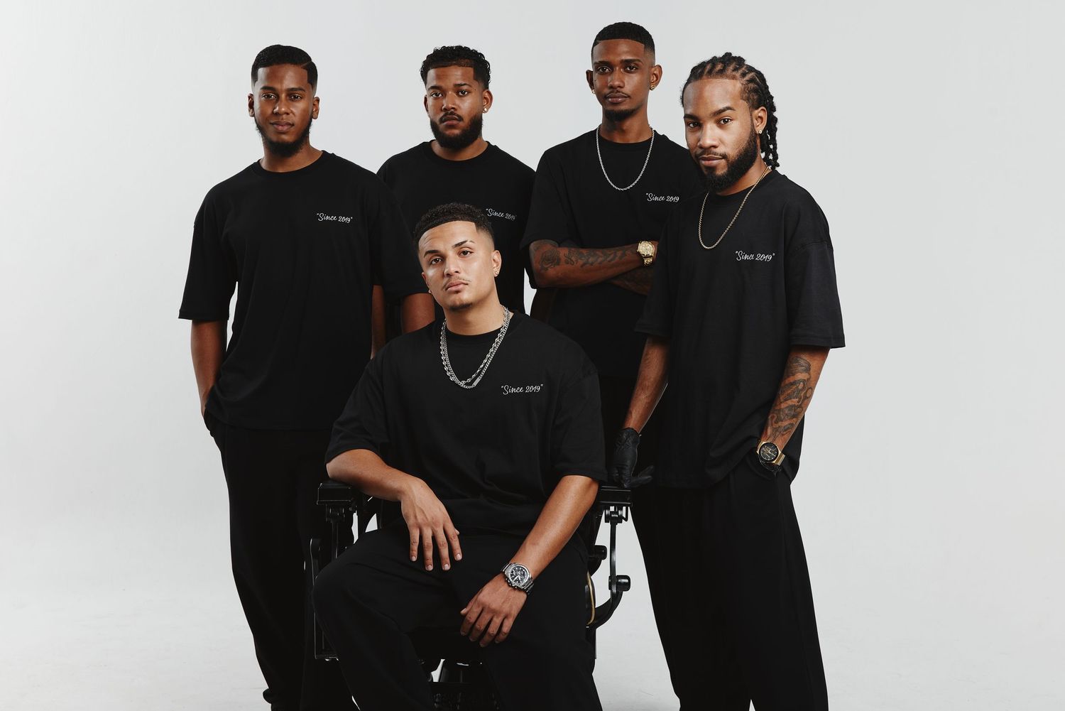 Group of men in black outfits pose together against white background for professional photoshoot.