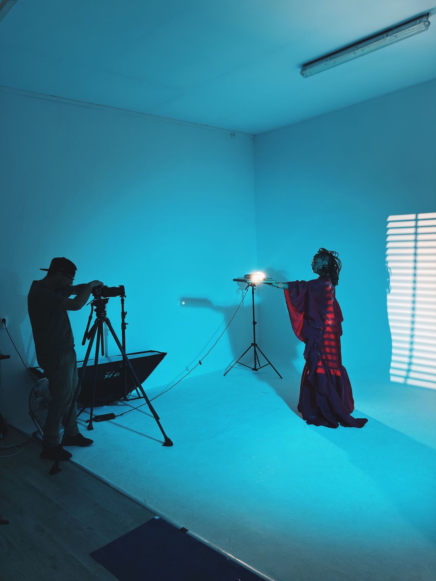 Photographer on tripod shoots model in colorful dress under blue studio lighting with softbox setup.