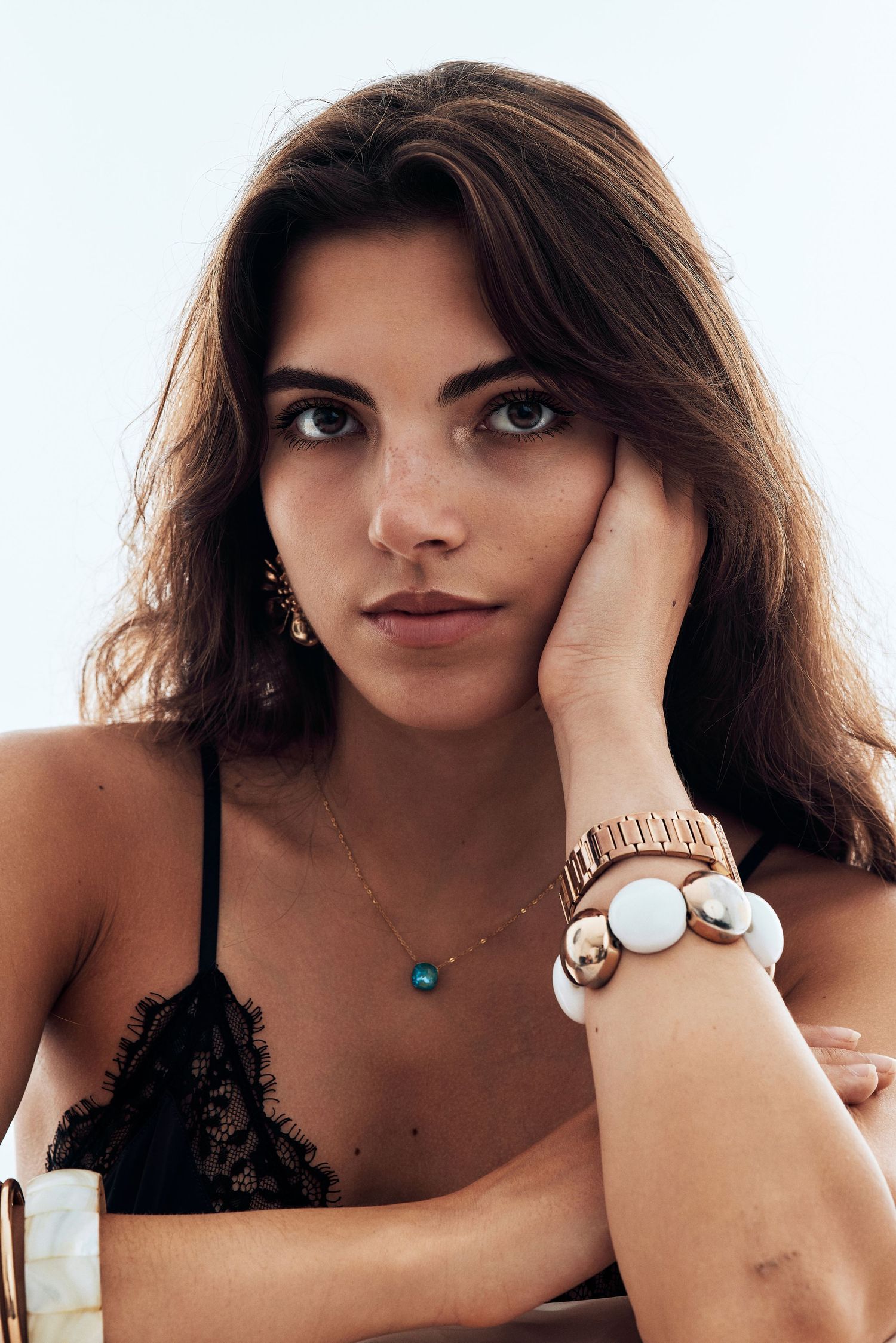 Woman wearing black lingerie poses seductively with jewelry and long brown hair against light background.