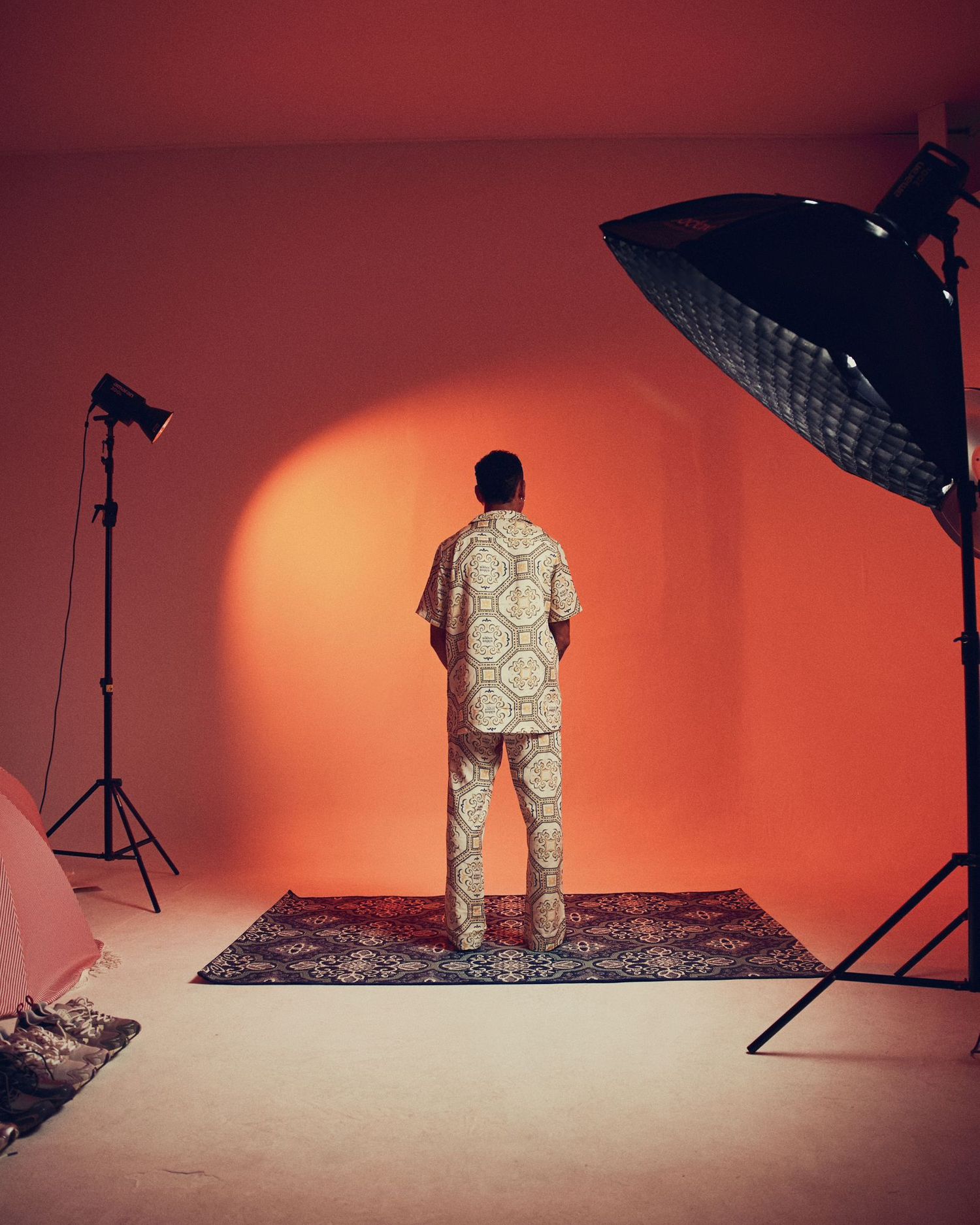 Fashion model posing on rug against orange backdrop with professional lighting setup.