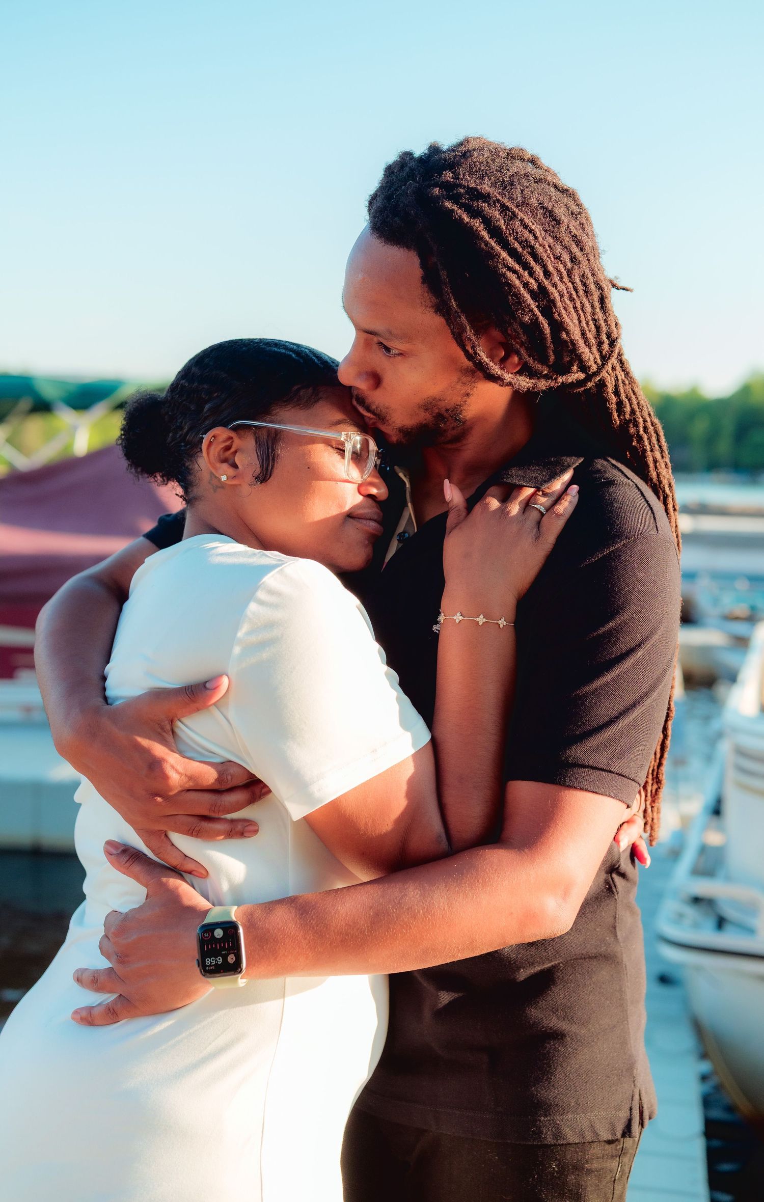A couple shares a tender kiss and embrace on a marina dock during golden hour, dressed in white and black outfits.