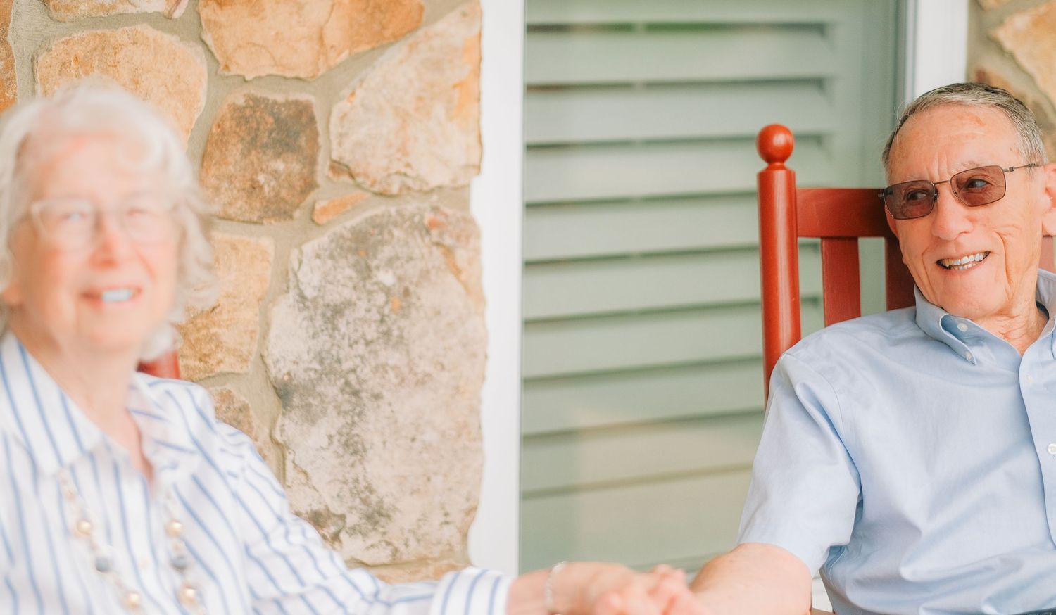 Elderly couple smiling and holding hands on a porch with a stone wall and green shutters.