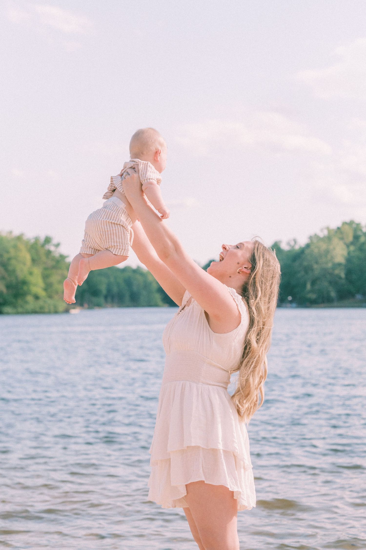 A joyful mother in a white dress lifts her baby high near a serene lake surrounded by lush green trees.