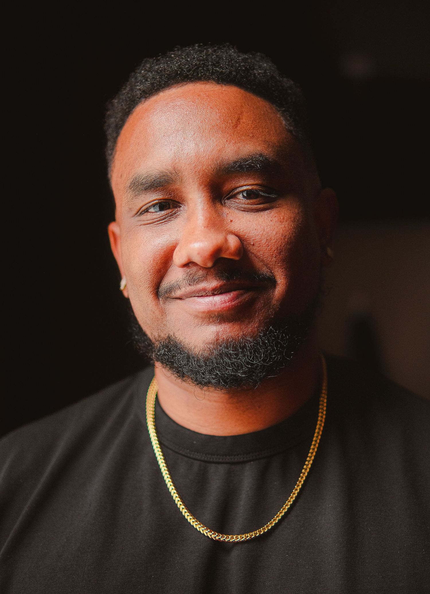 Smiling young Black man with beard wearing black t-shirt and gold chain necklace against dark background.