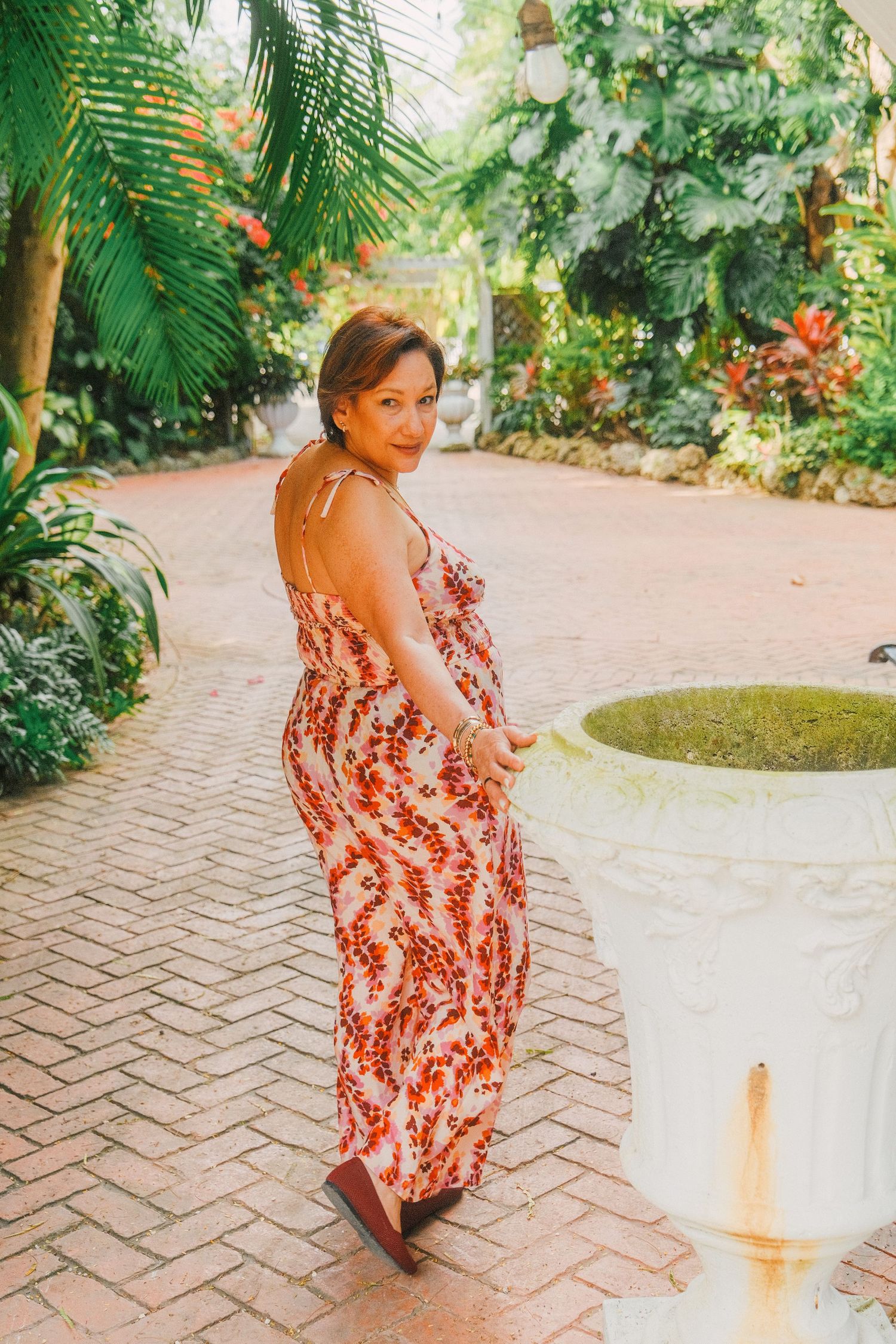 A person in a red floral dress walks along a brick pathway in a lush tropical garden setting.