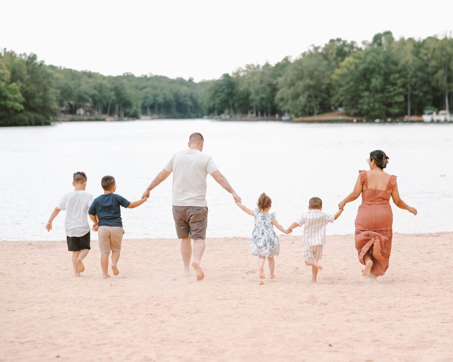 Family of six walking hand-in-hand on sandy beach toward calm lake surrounded by lush green trees.