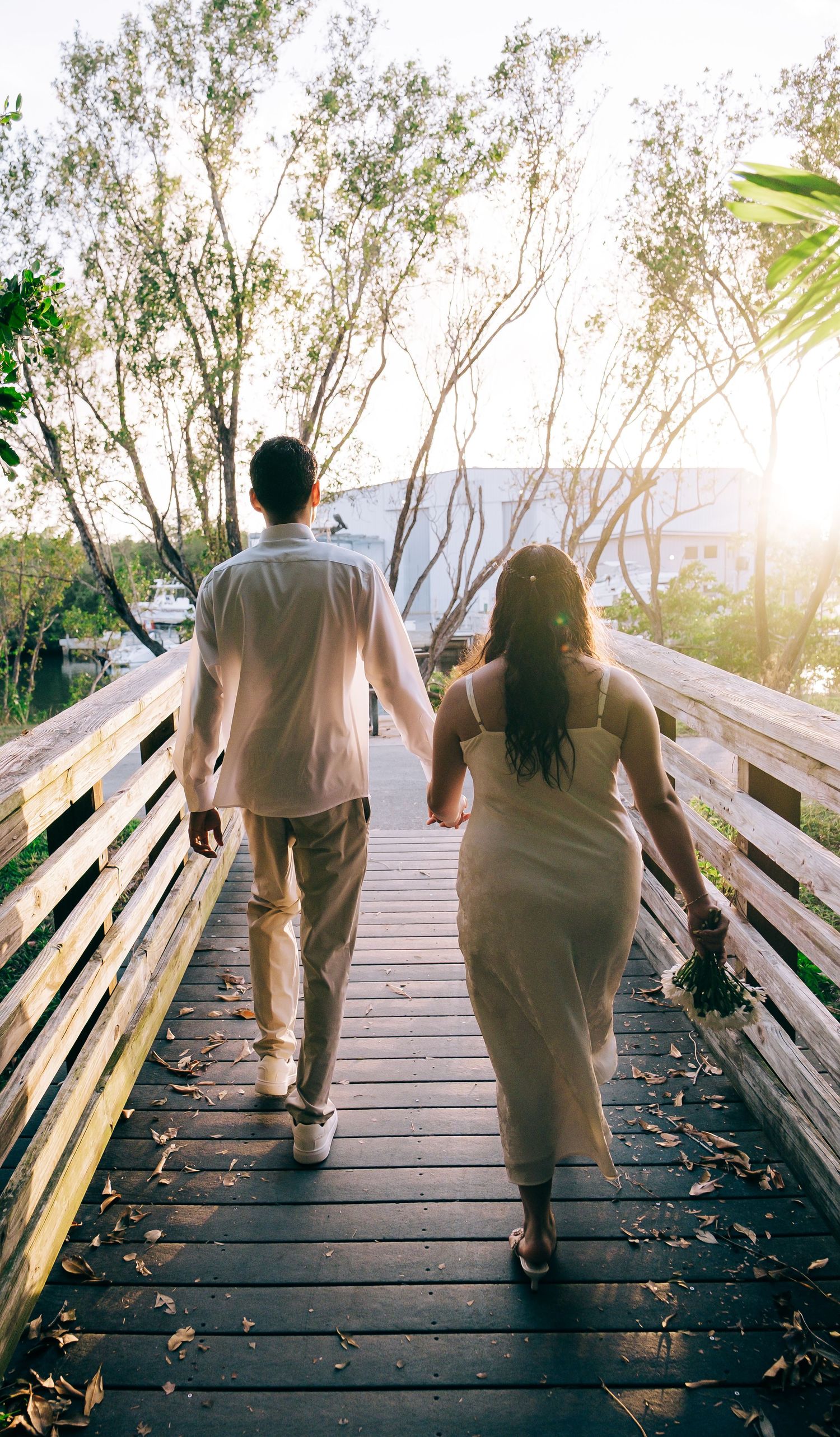 A couple walks hand in hand on a wooden boardwalk during sunset surrounded by trees and natural light.