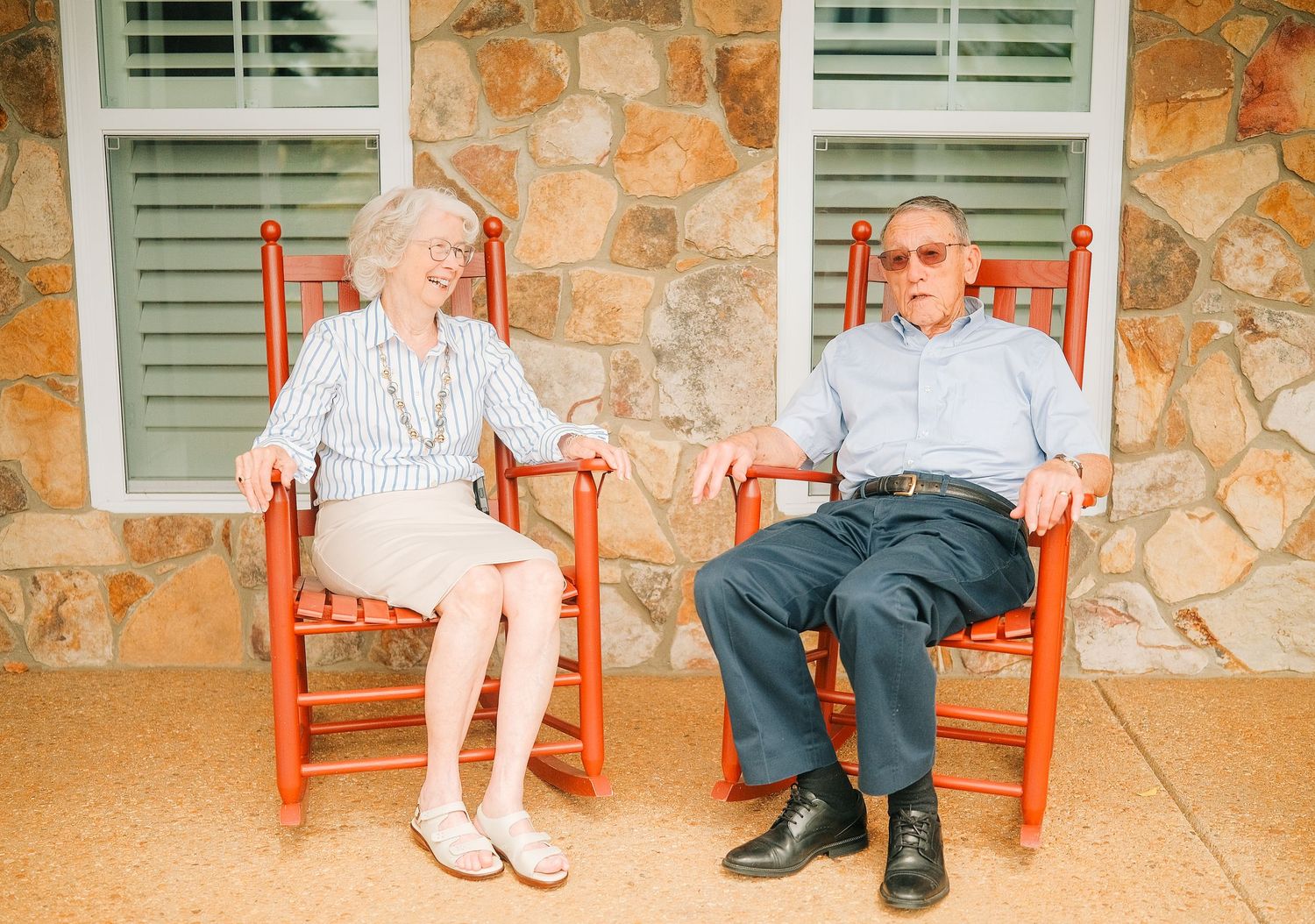 Elderly couple holding hands while sitting in red rocking chairs on a stone porch.