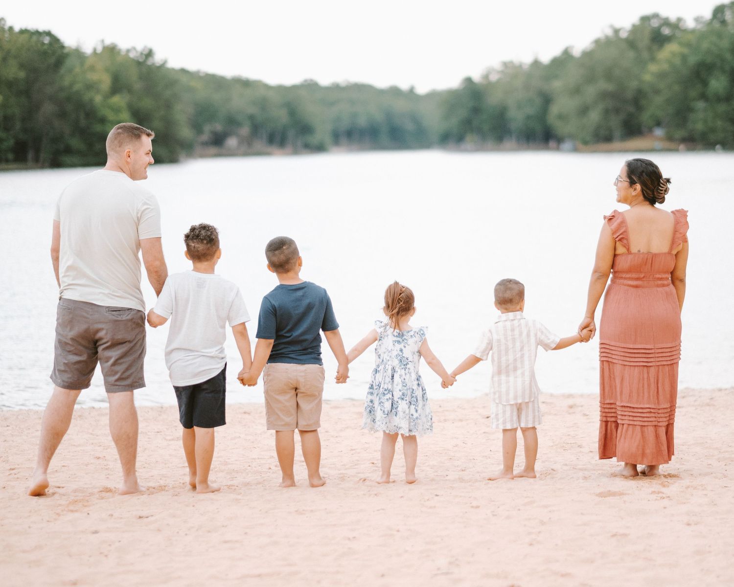 Family of seven holding hands on sandy beach shore with serene lake and lush green trees in background.