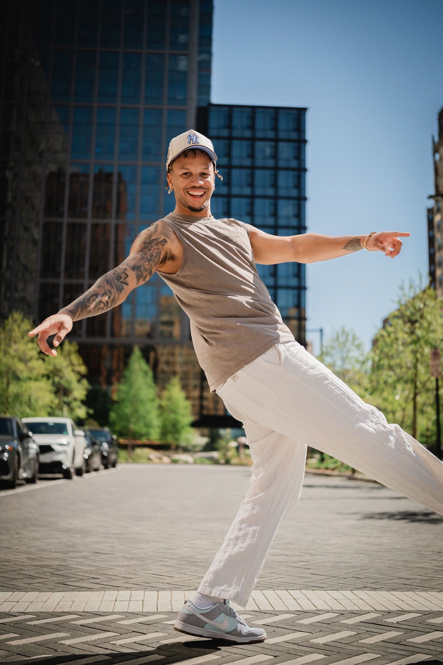 Smiling tattooed man in white pants and cap dancing outdoors in urban setting with modern glass building.