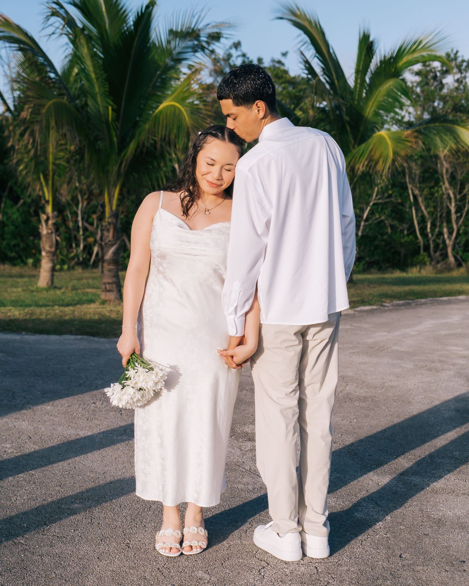 A couple in white outfits holds hands against a backdrop of palm trees on a sunny tropical day.