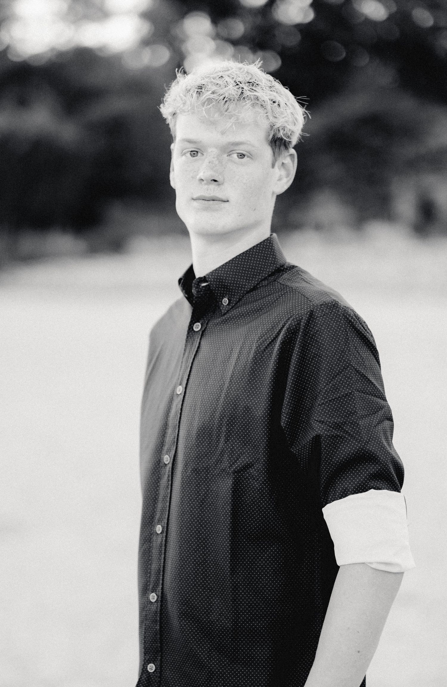 Black and white portrait of a young man with curly blonde hair wearing a button-up shirt outdoors.