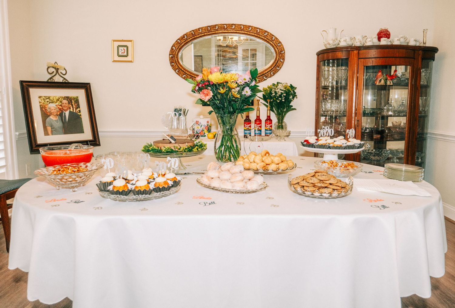 Elegant party buffet table with white tablecloth, assorted cookies, floral centerpiece, and framed photo in a decorated room.