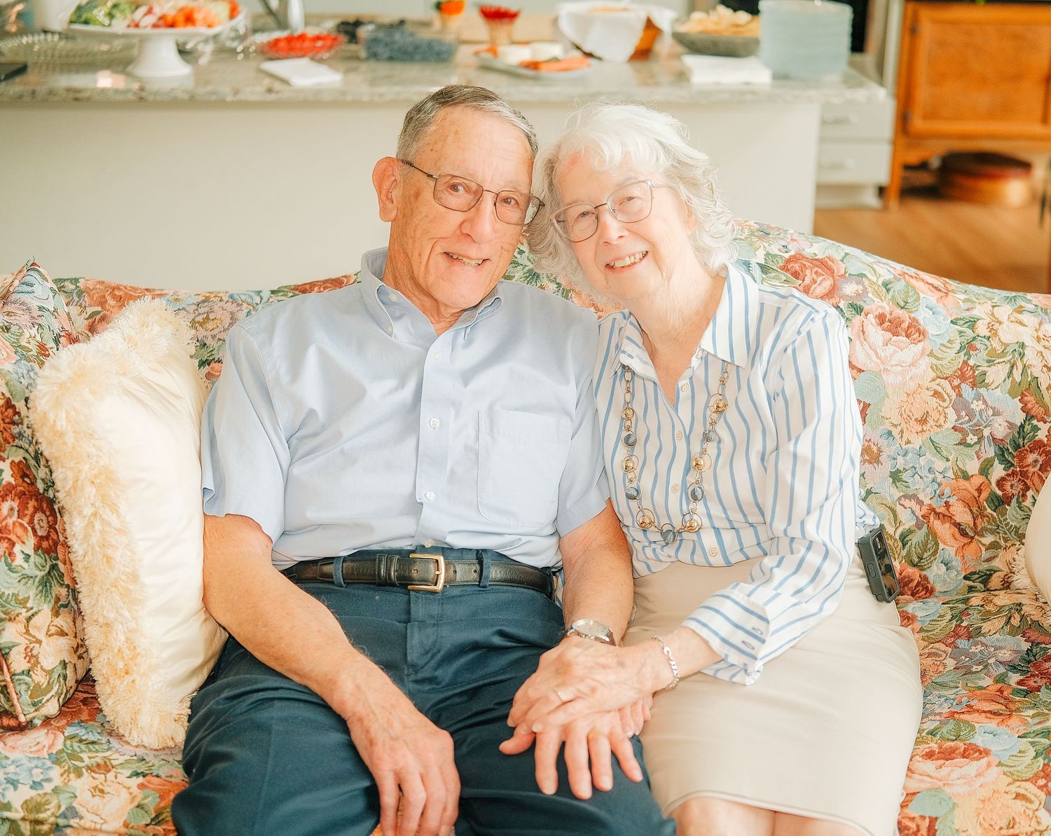 Elderly couple smiling and sitting together on a floral sofa in a bright, cozy living room.
