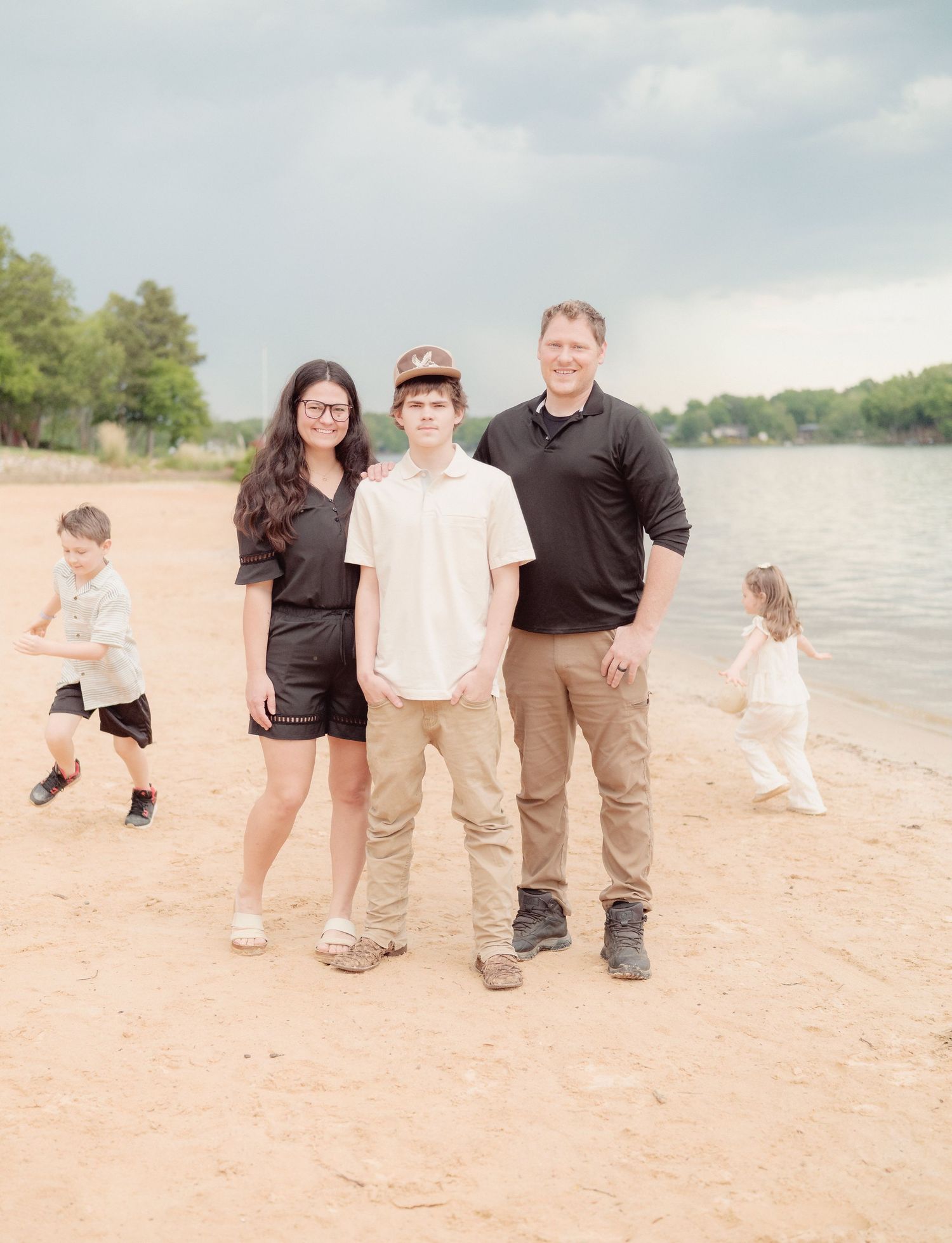 Family of four poses on sandy beach with two young children playing behind them, trees and cloudy sky in background.