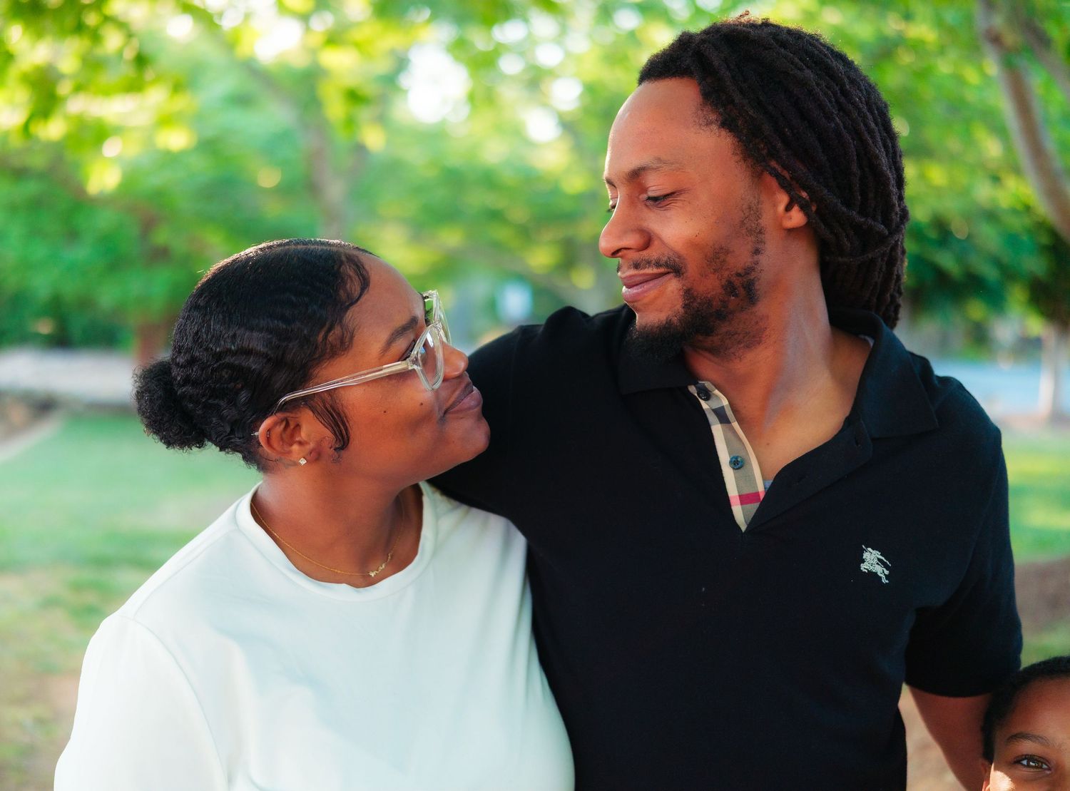 A joyful Black couple smiling at each other outdoors, surrounded by lush green trees on a sunny day.
