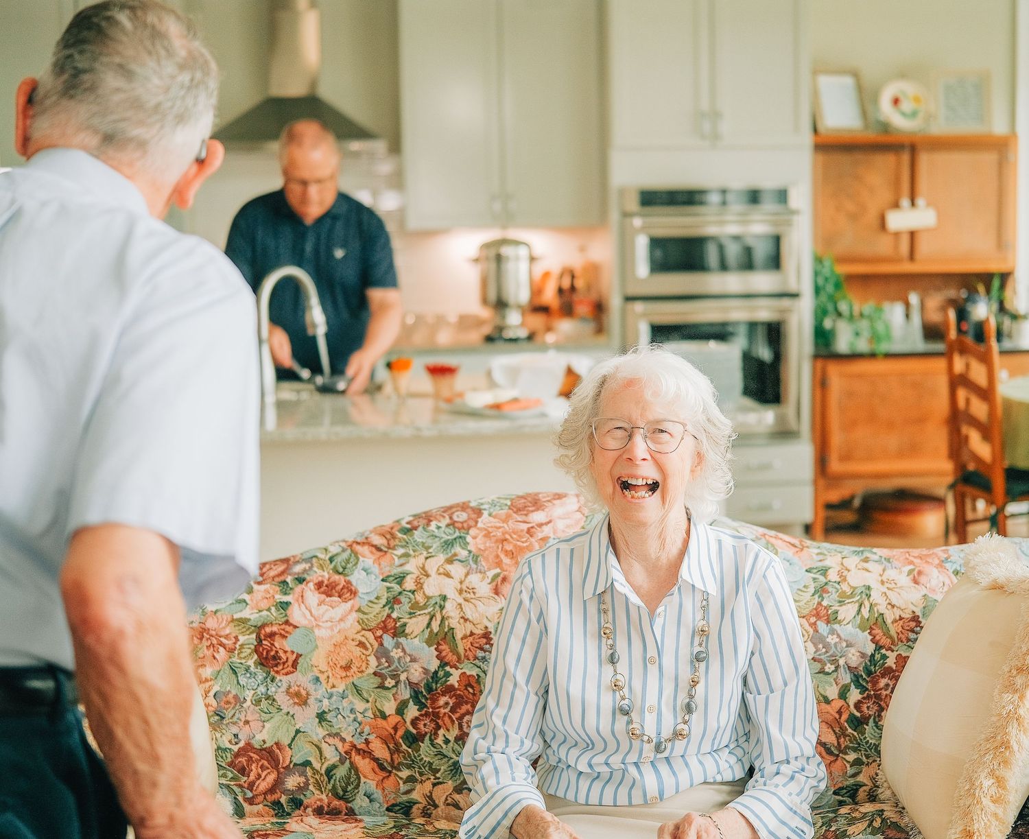 Elderly woman laughing joyfully on floral sofa with two men in bright home kitchen setting.
