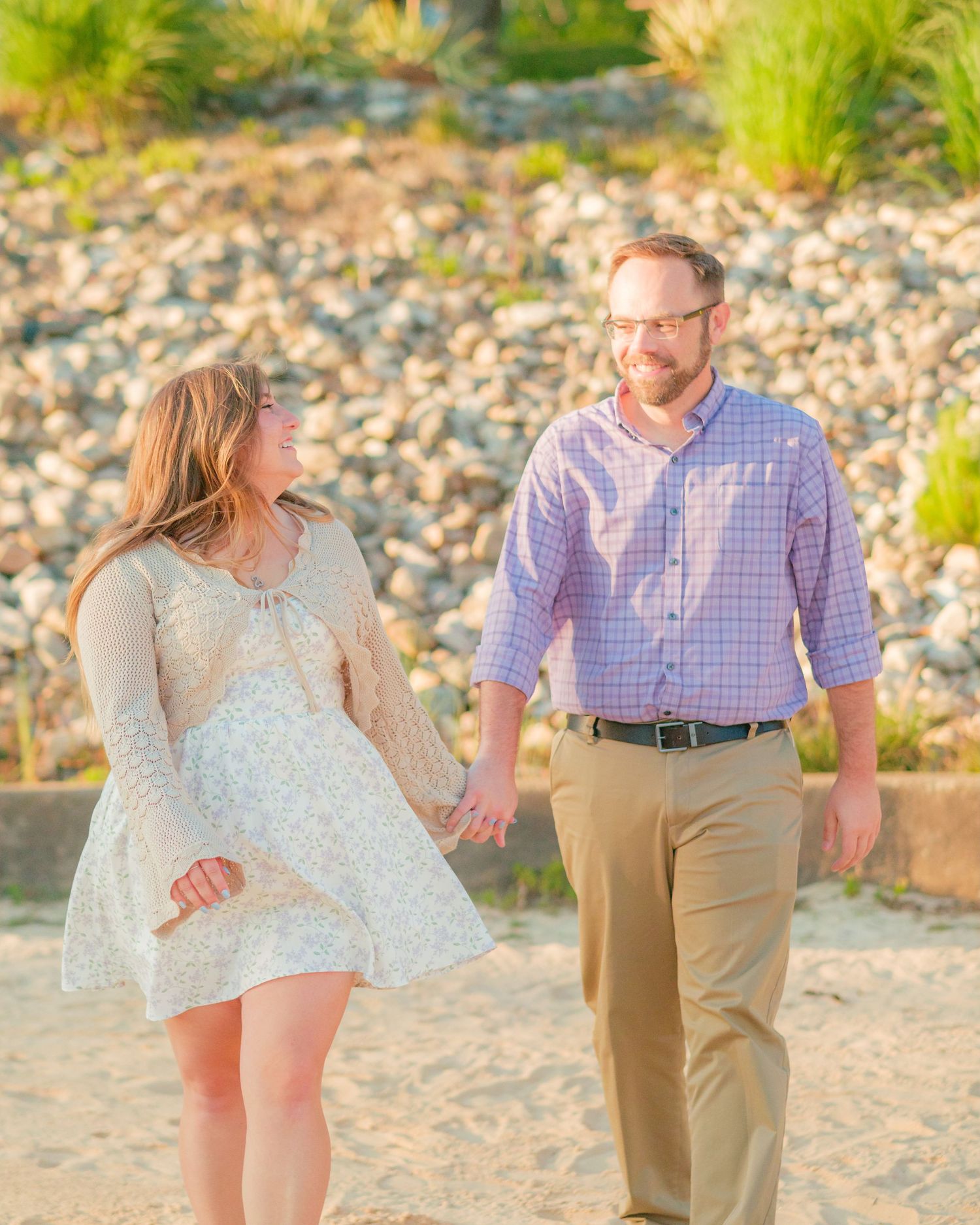 Couple holding hands and smiling at each other on a sandy beach with rocky shoreline backdrop in warm golden light.