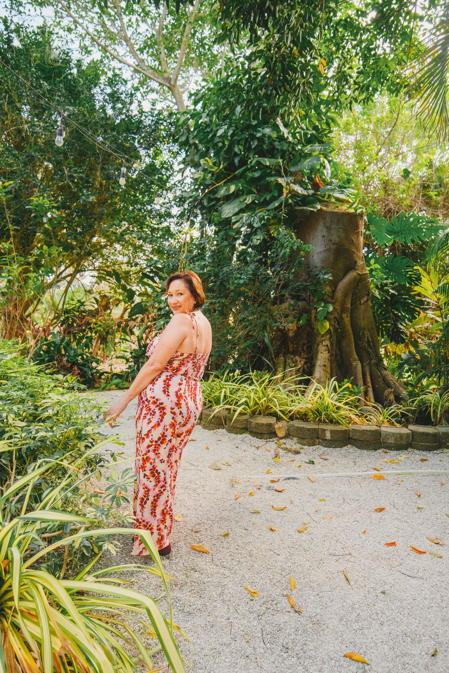 Person in a flowing red and white floral dress walks along a sandy tropical garden path surrounded by lush greenery.