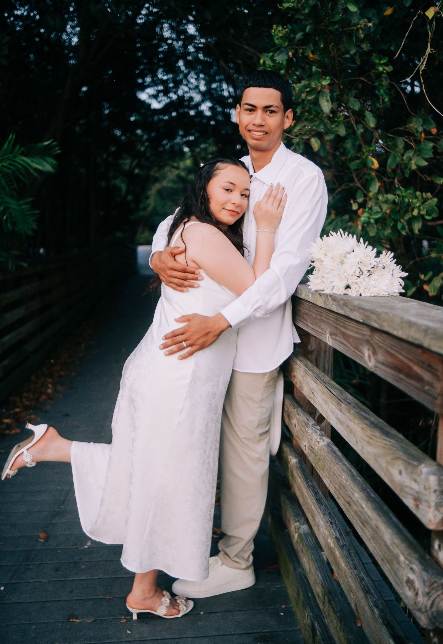 Couple in white clothing sharing tender moments on a wooden boardwalk surrounded by greenery.