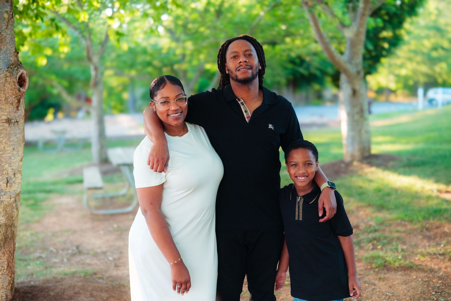 A family of three smiling outdoors in a park, with a woman in white, a tall man, and a young boy.
