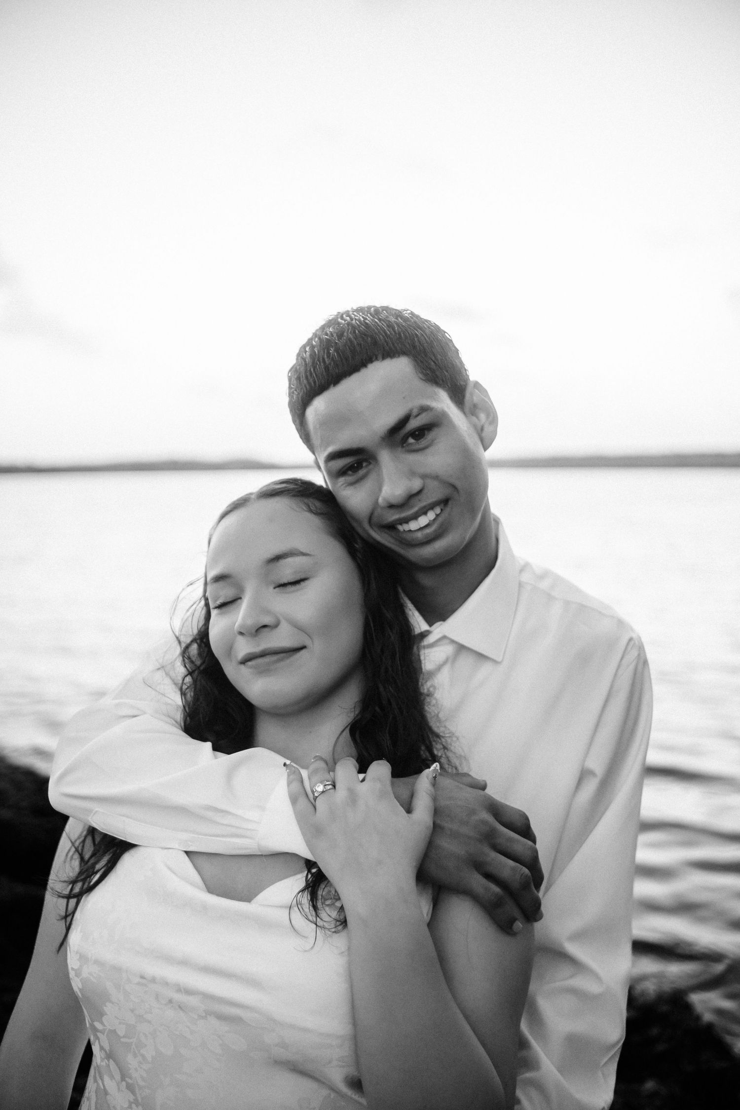 Romantic black and white portrait of a couple embracing by the water at sunset.
