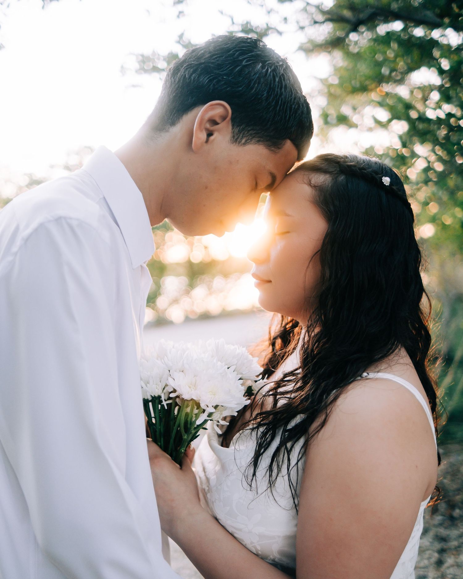 Intimate couple in white embracing outdoors with soft sunlight filtering through trees in the background.