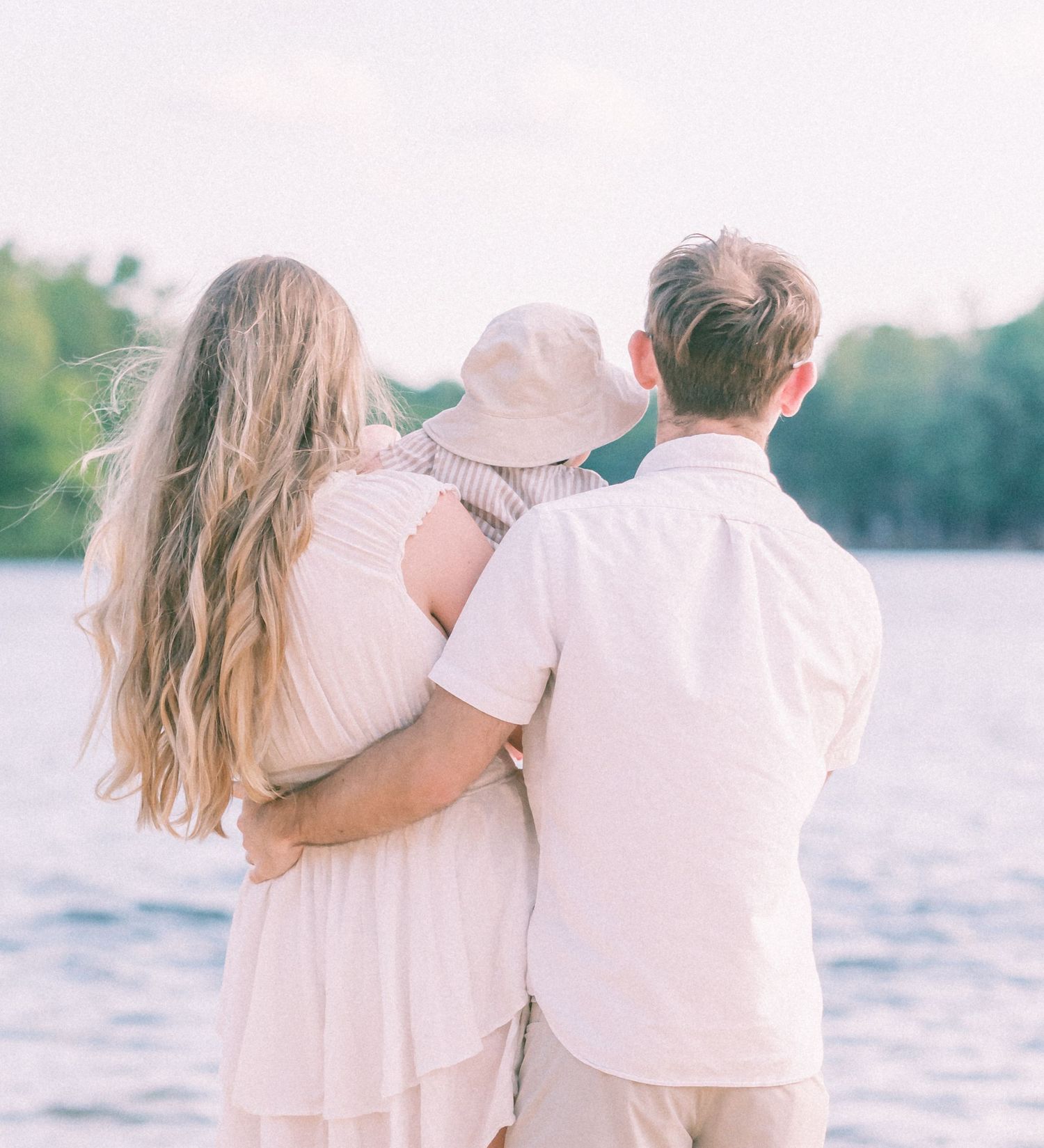 A family of three with a young child in a hat stands embracing by a serene lake, viewed from behind.
