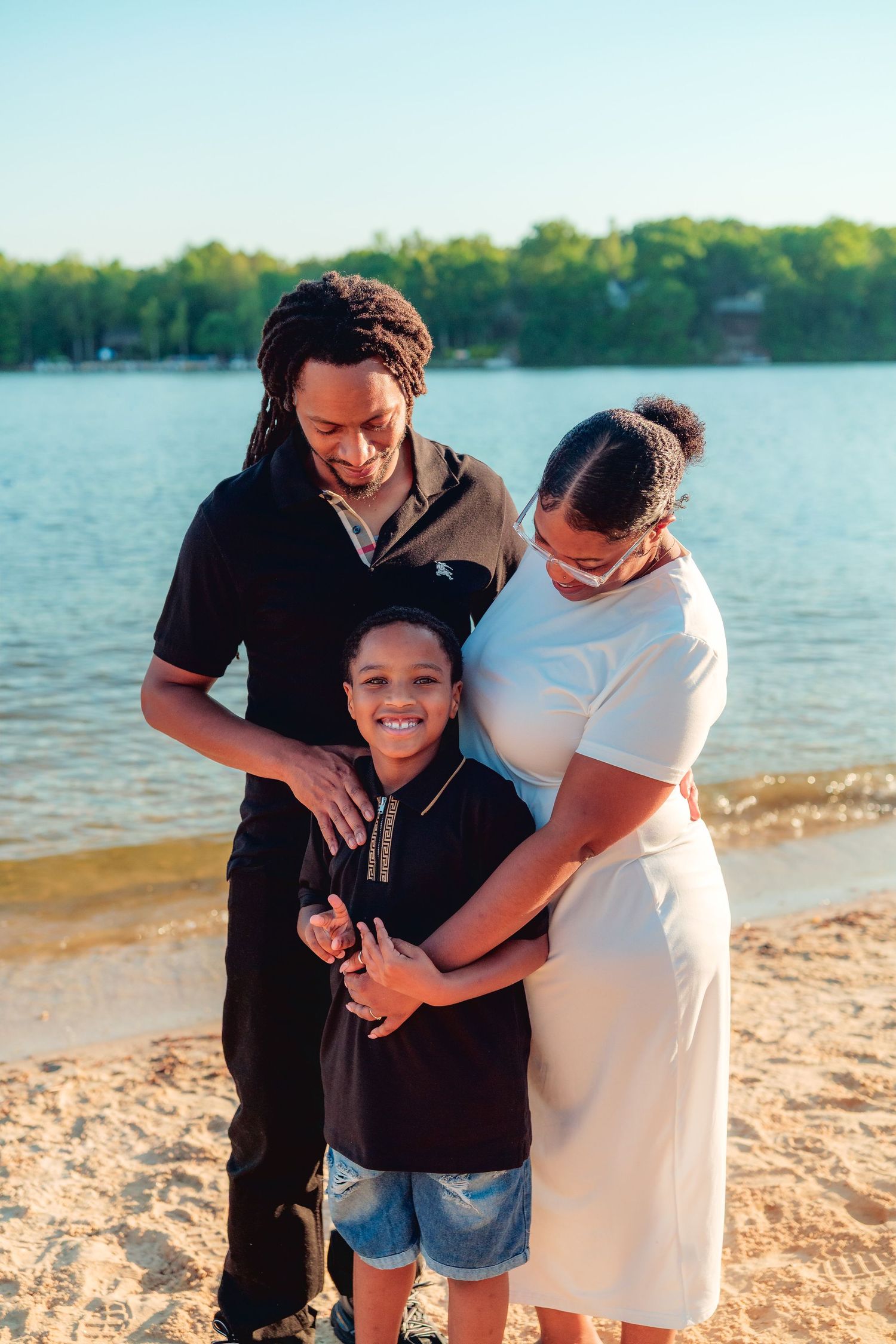 A happy family of three poses on a sandy beach by a lake, with parents embracing their smiling young son.