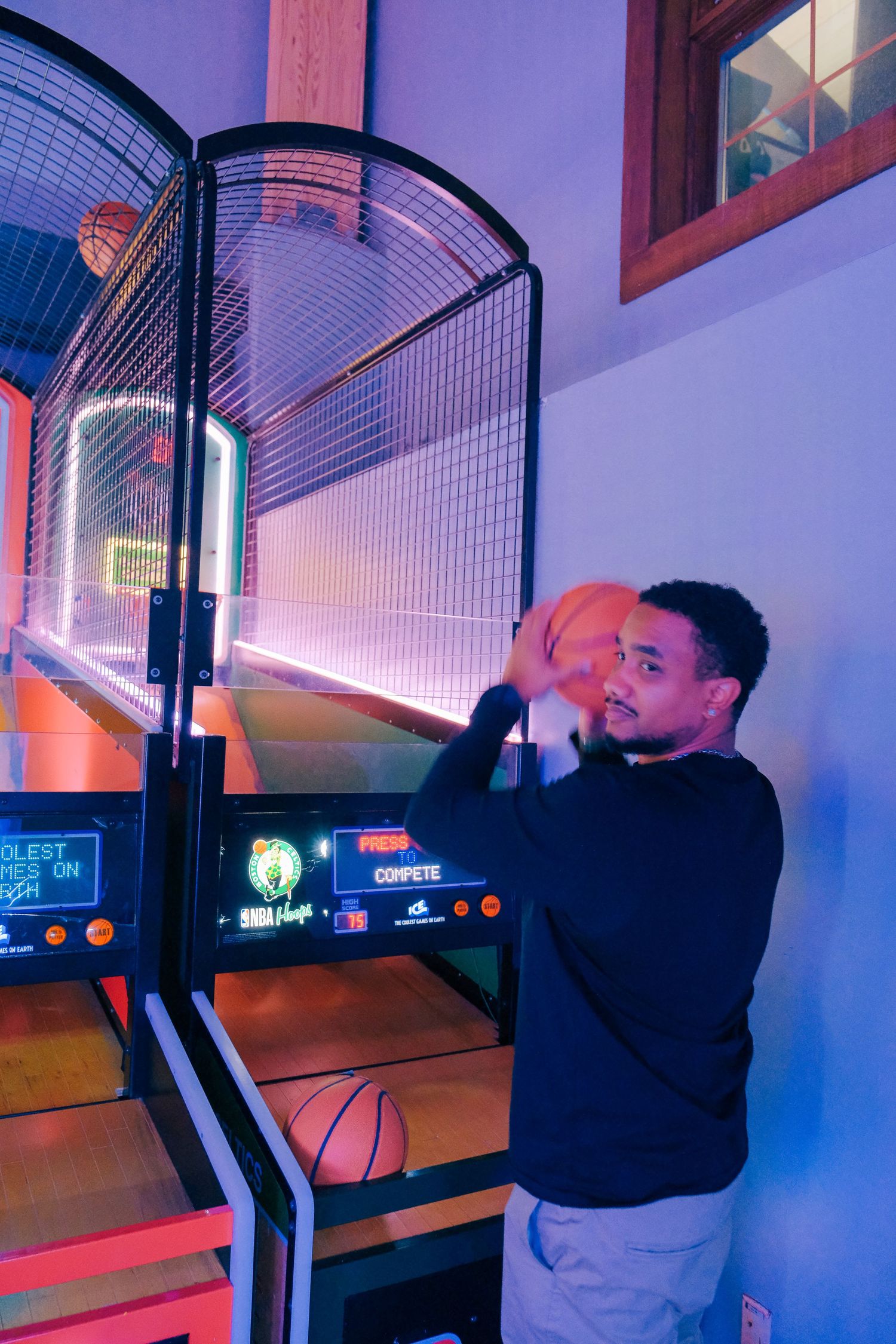 Person playing arcade basketball game in dimly lit room with neon blue and pink lighting.
