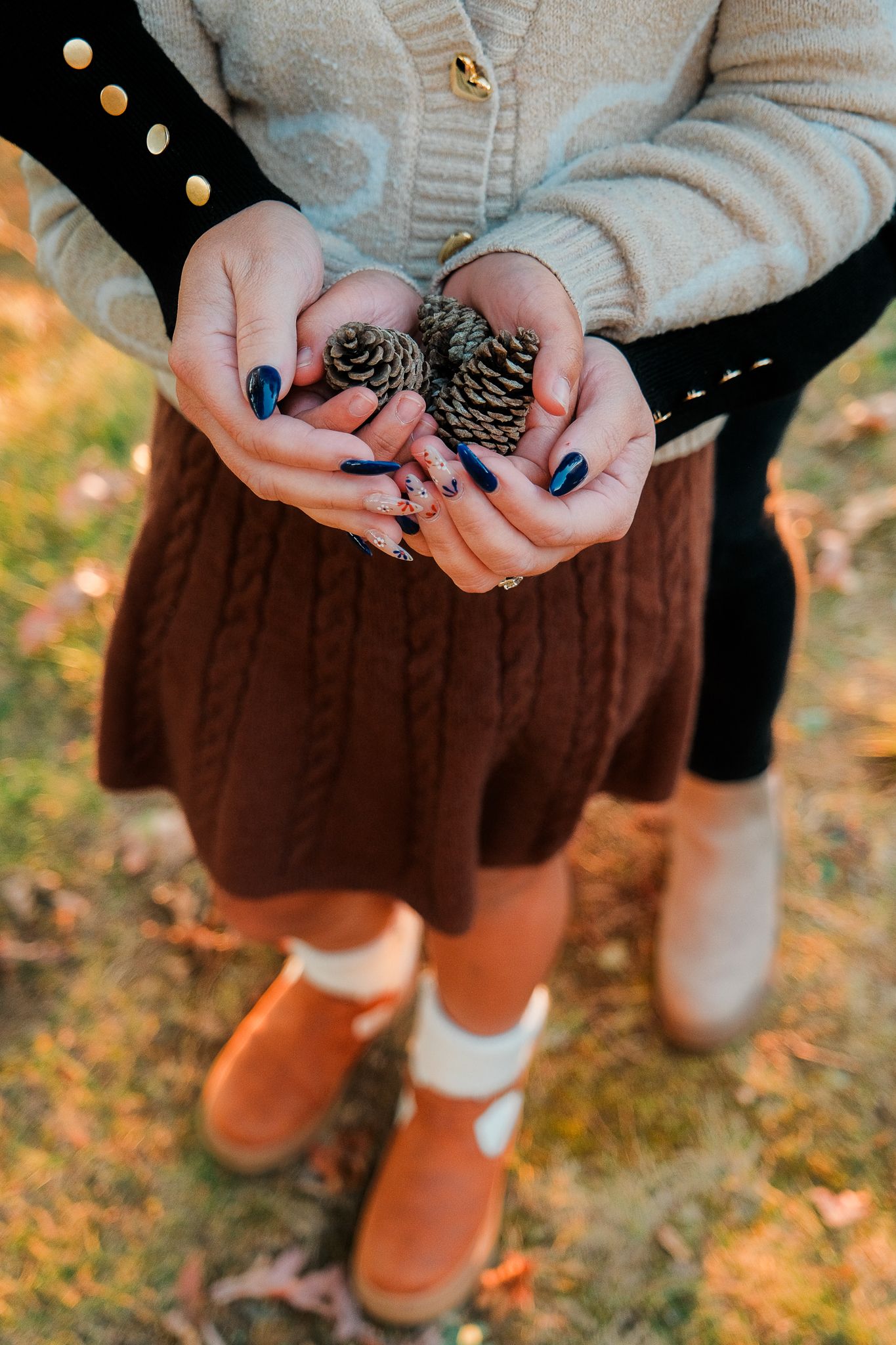 Close-up of hands holding pine cones while wearing a beige sweater and brown skirt.