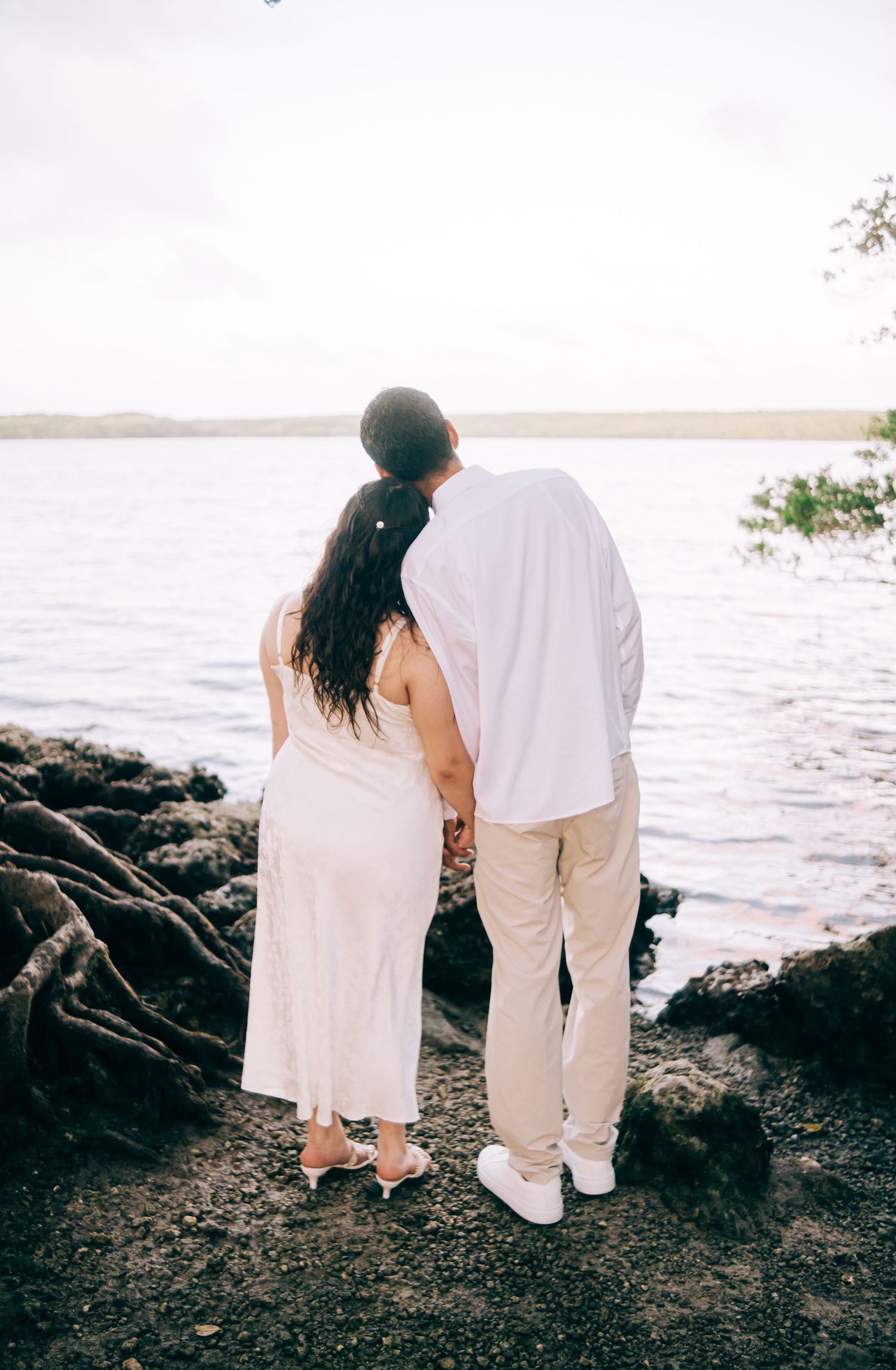 Couple holding hands facing a serene waterfront, woman in white dress and man in white shirt and khaki pants.