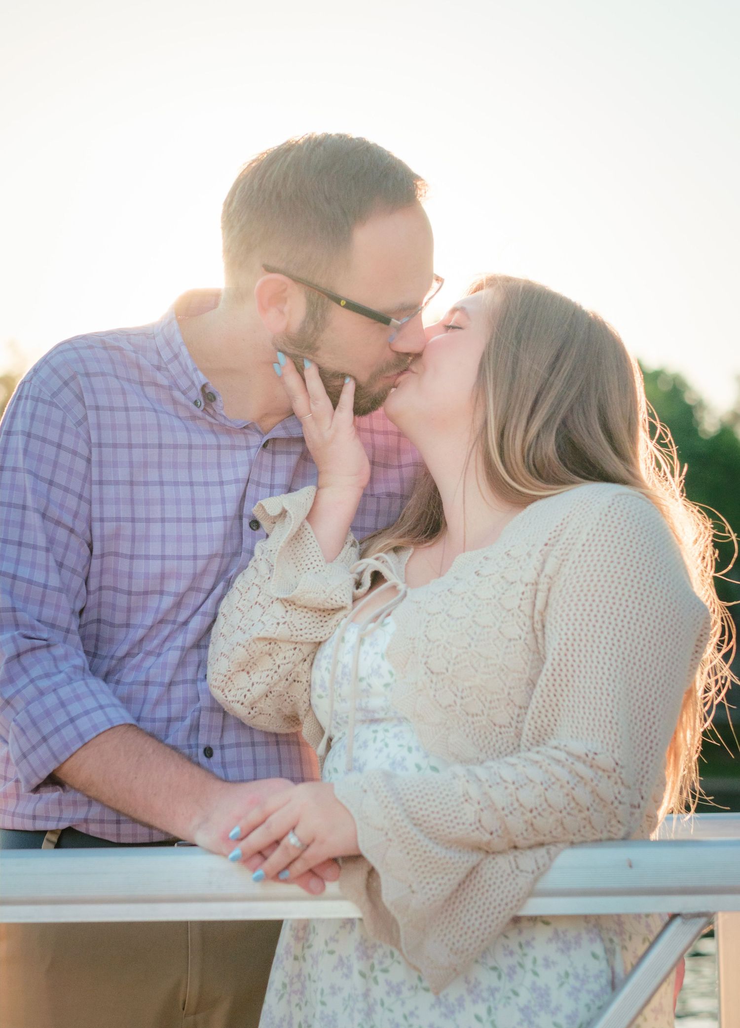 Couple sharing a romantic kiss outdoors in golden hour sunlight, woman in lace dress and man in plaid shirt.