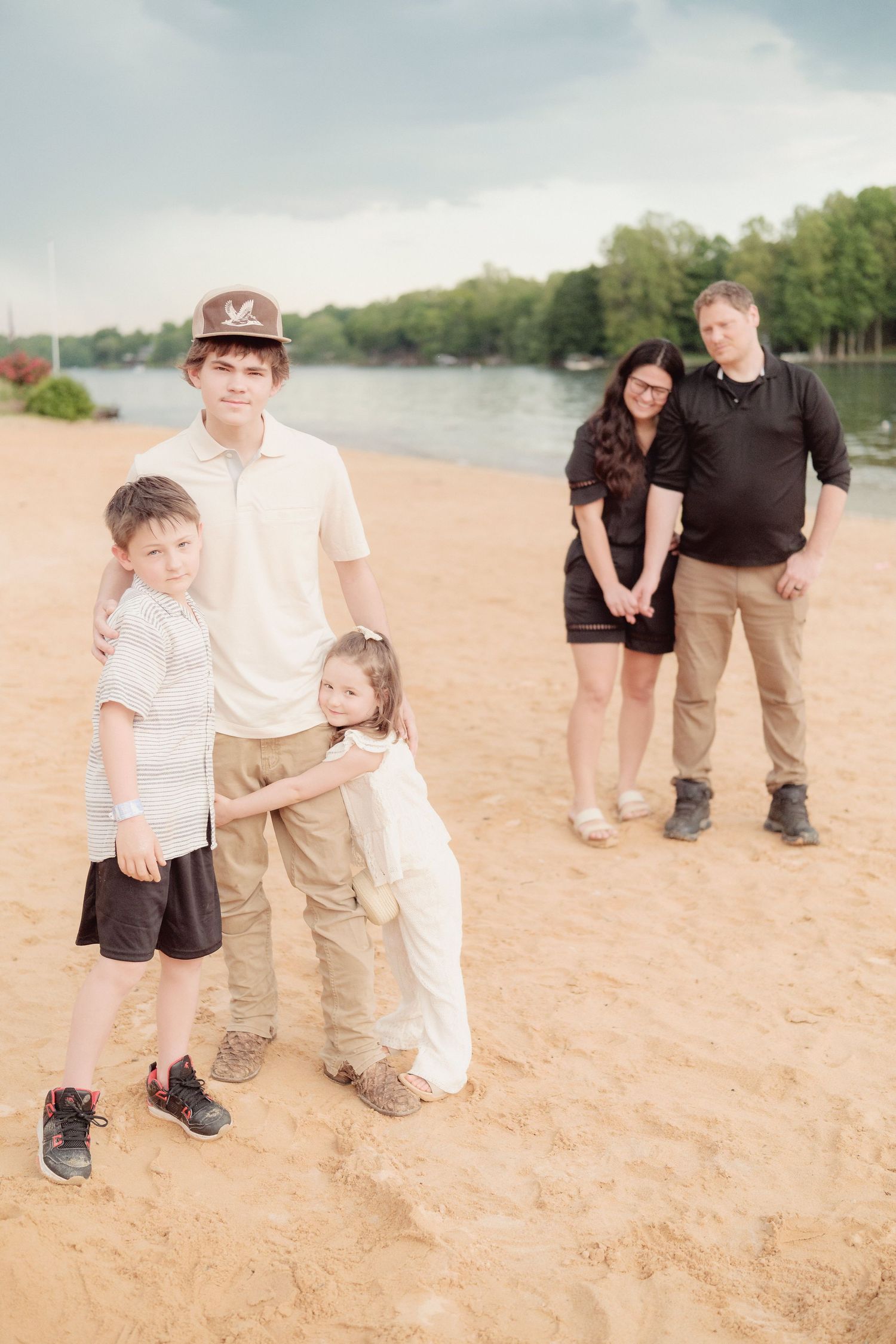 Family of five posing on sandy beach near lake, three children in foreground with parents standing behind them.