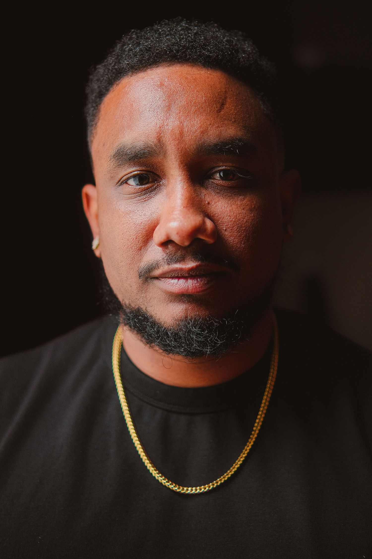 A young Black man with a goatee wearing a black t-shirt and gold chain necklace poses against a dark background.