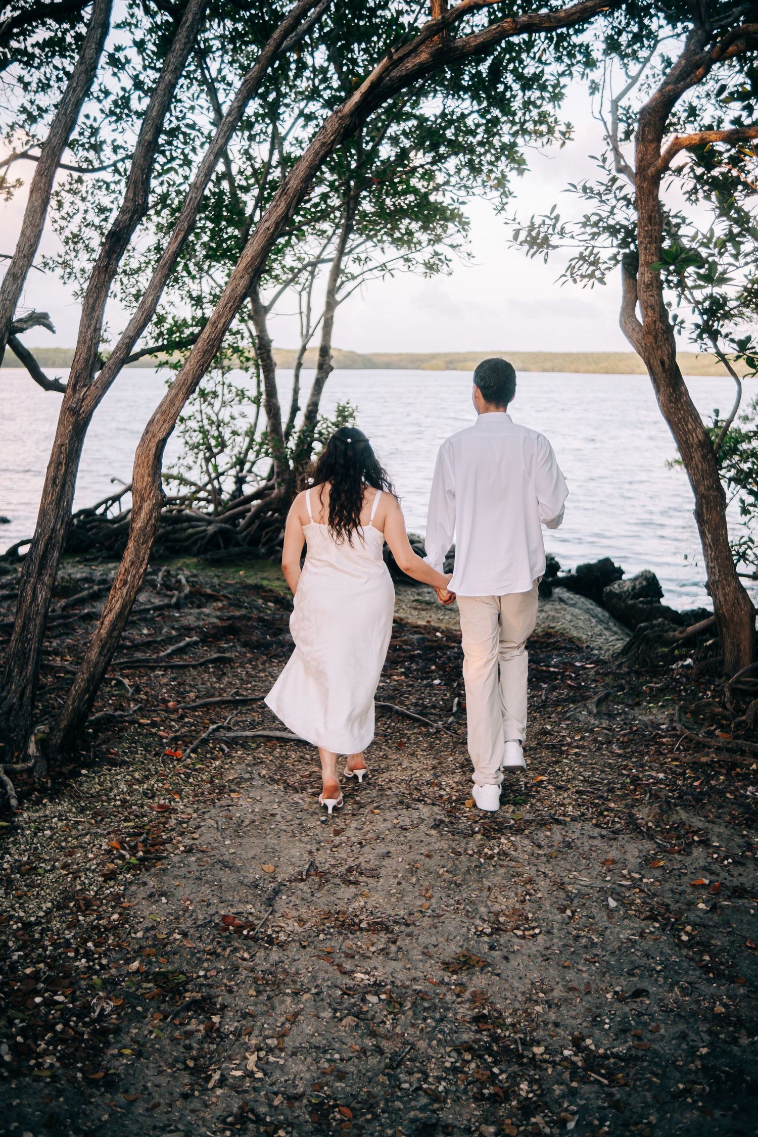 A couple in white clothing walks along a rocky shoreline beneath trees at sunset.