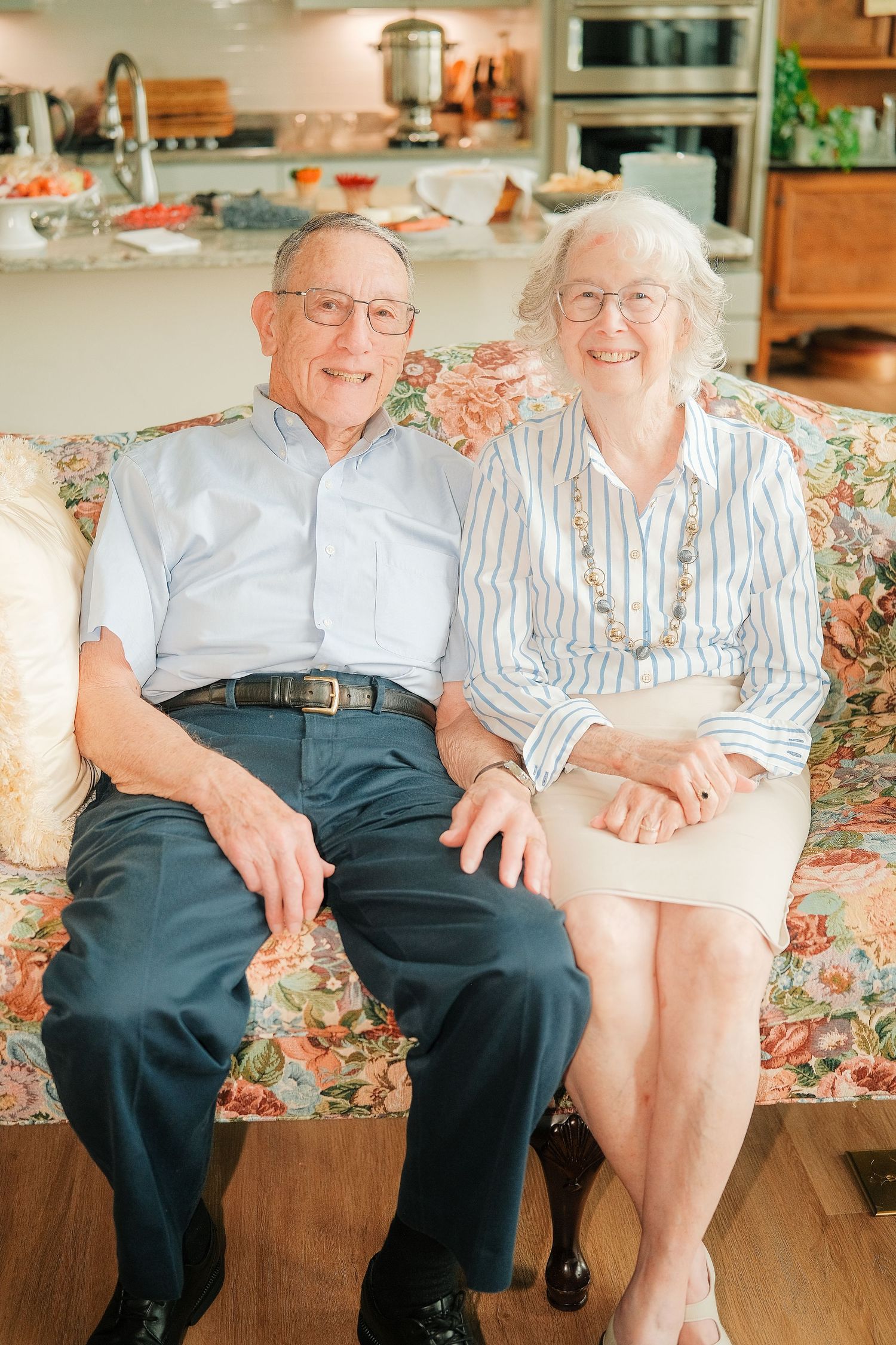 Elderly couple smiling and holding hands on a floral sofa in a bright home setting.