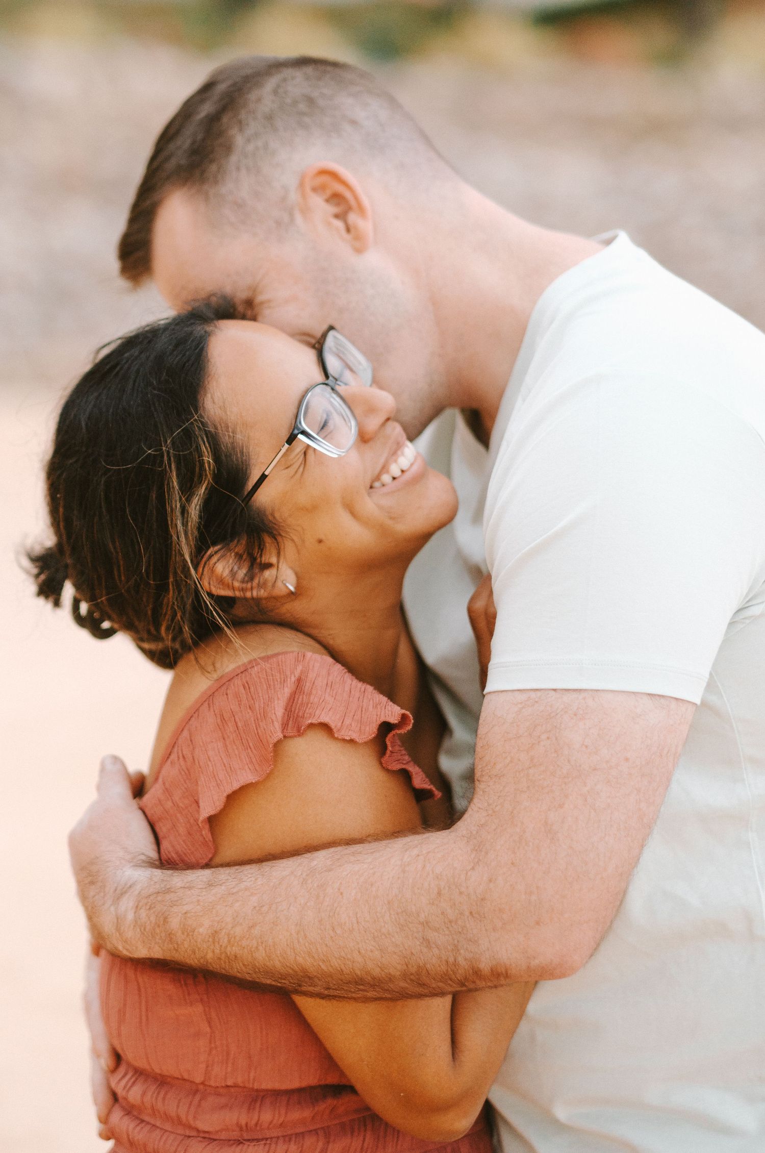 A couple embraces warmly outdoors, woman in pink dress smiling up at man in white t-shirt kissing her forehead.