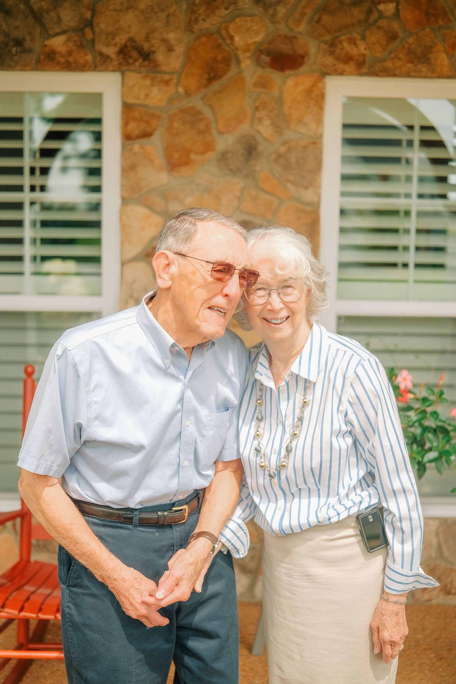 Elderly couple smiling and leaning together outdoors in front of a stone house, wearing casual summer attire.