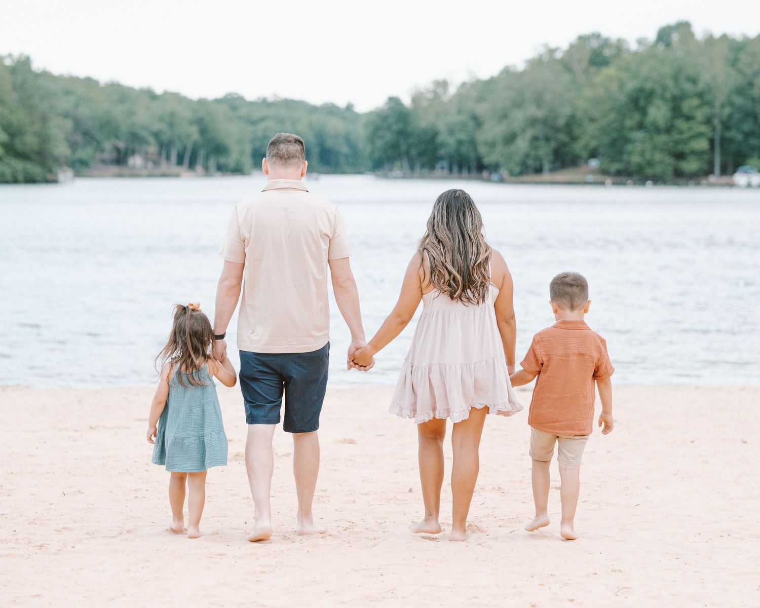 Family of four holding hands walking toward a lake on a sandy beach, viewed from behind.